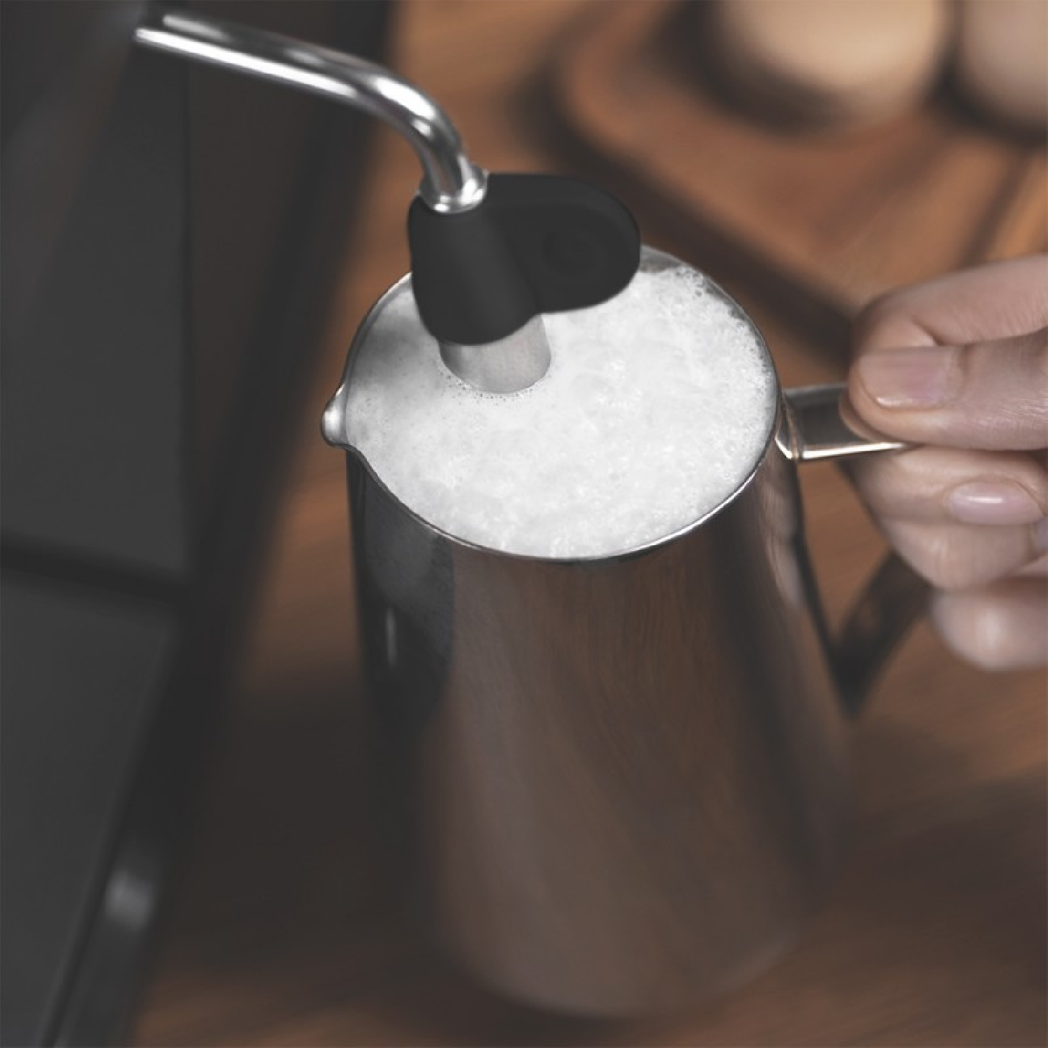 Dzbanek do spieniania mleka ze stali nierdzewnej z czarną dyszą, spieniający mleko. Ręka trzyma dzbanek.