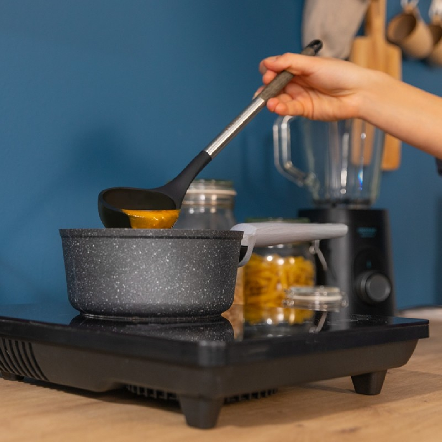 Una persona versa la zuppa da un mestolo nero in una pentola su un fornello elettrico, con oggetti da cucina sullo sfondo.