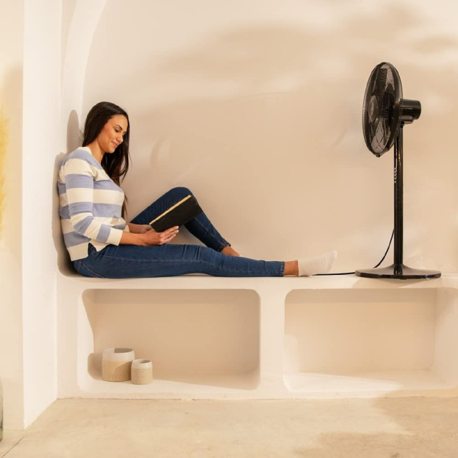 Kobieta siedzi na gzymsie i czyta. Obok niej stoi czarny wentylator.