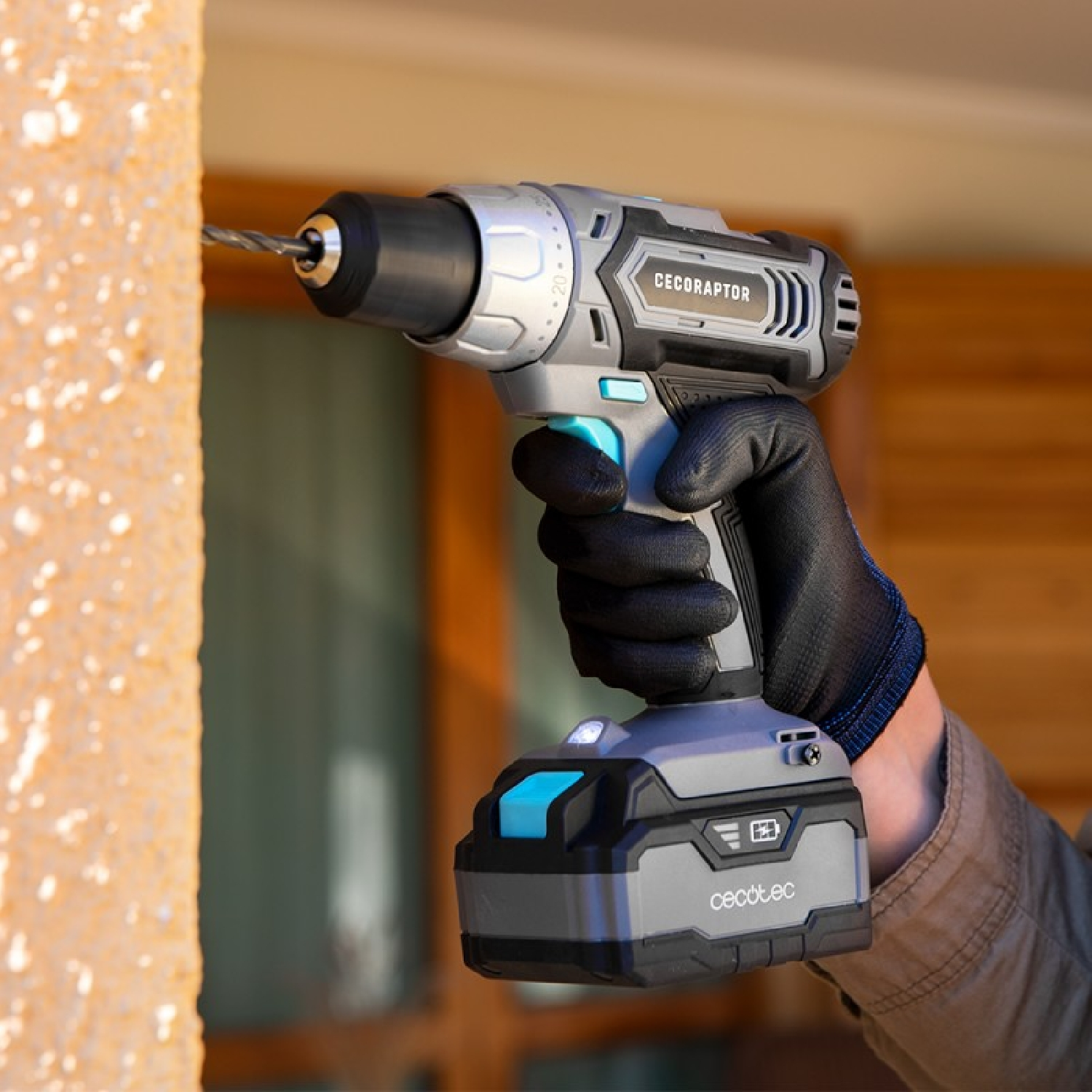 Una persona usando un taladro Cecotec gris y negro en una pared, usando un guante negro.