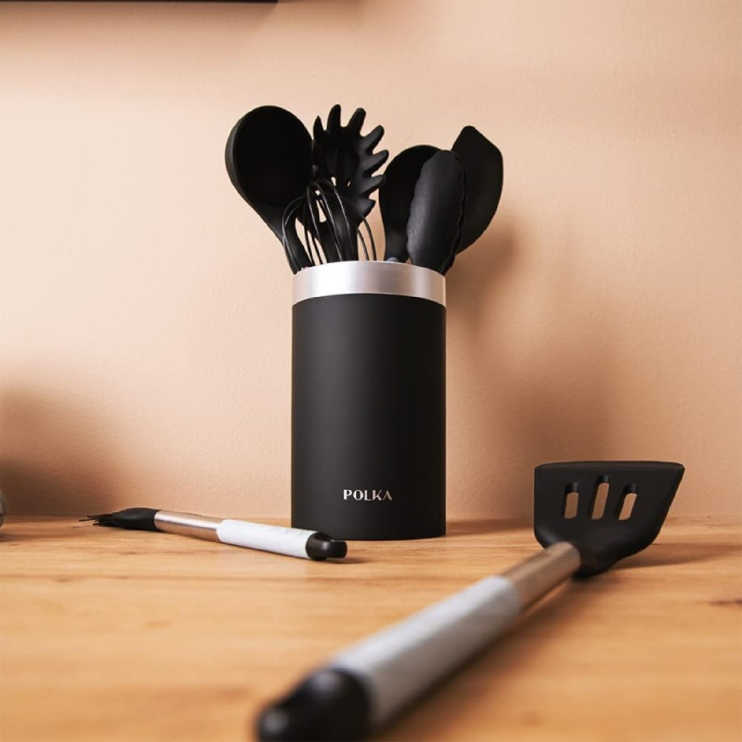 Black kitchen utensils in a holder, and two utensils in front. All on a wooden table with a beige background.