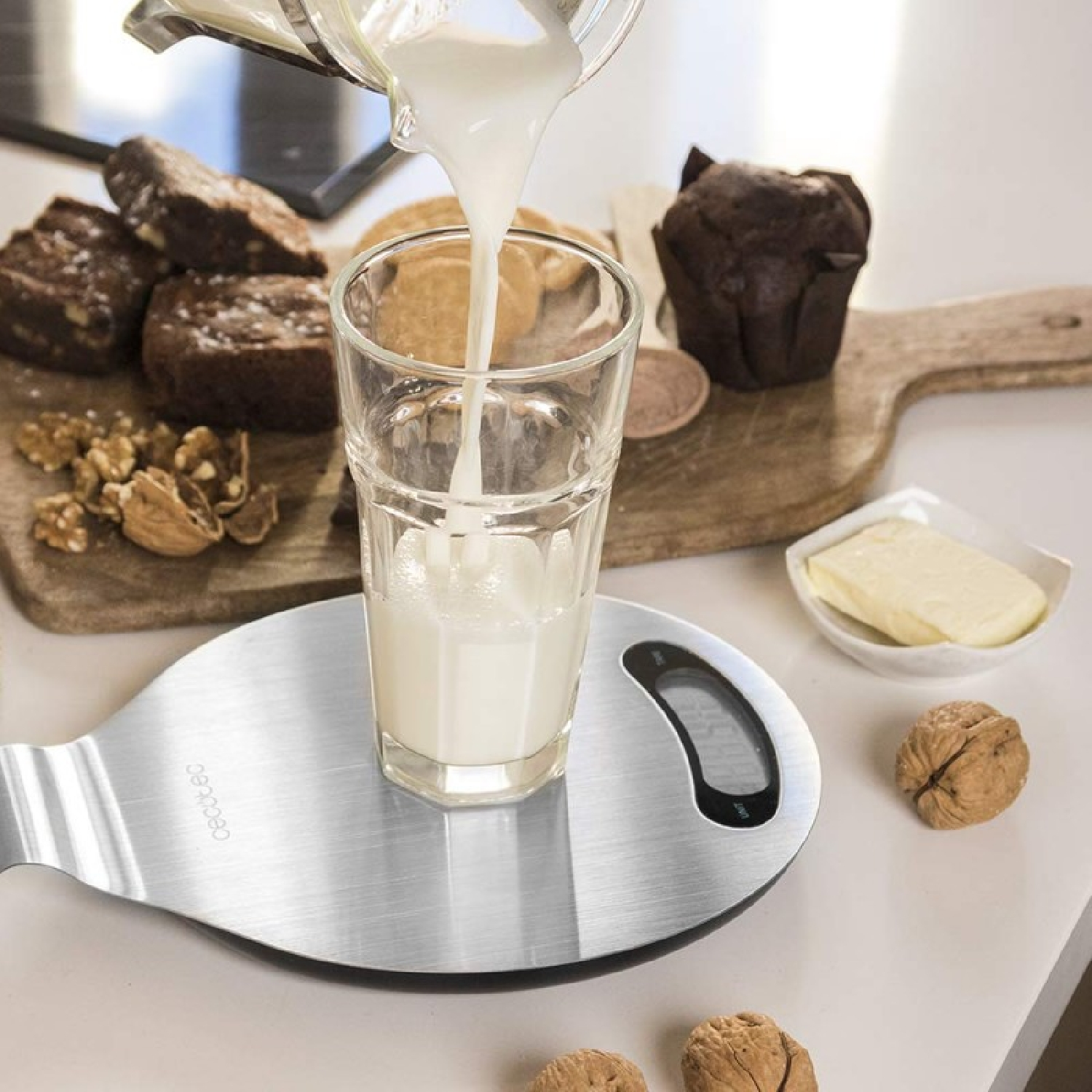 Mleko wlewa się do szklanki na wadze. Brownie, masło i orzechy włoskie na stole.