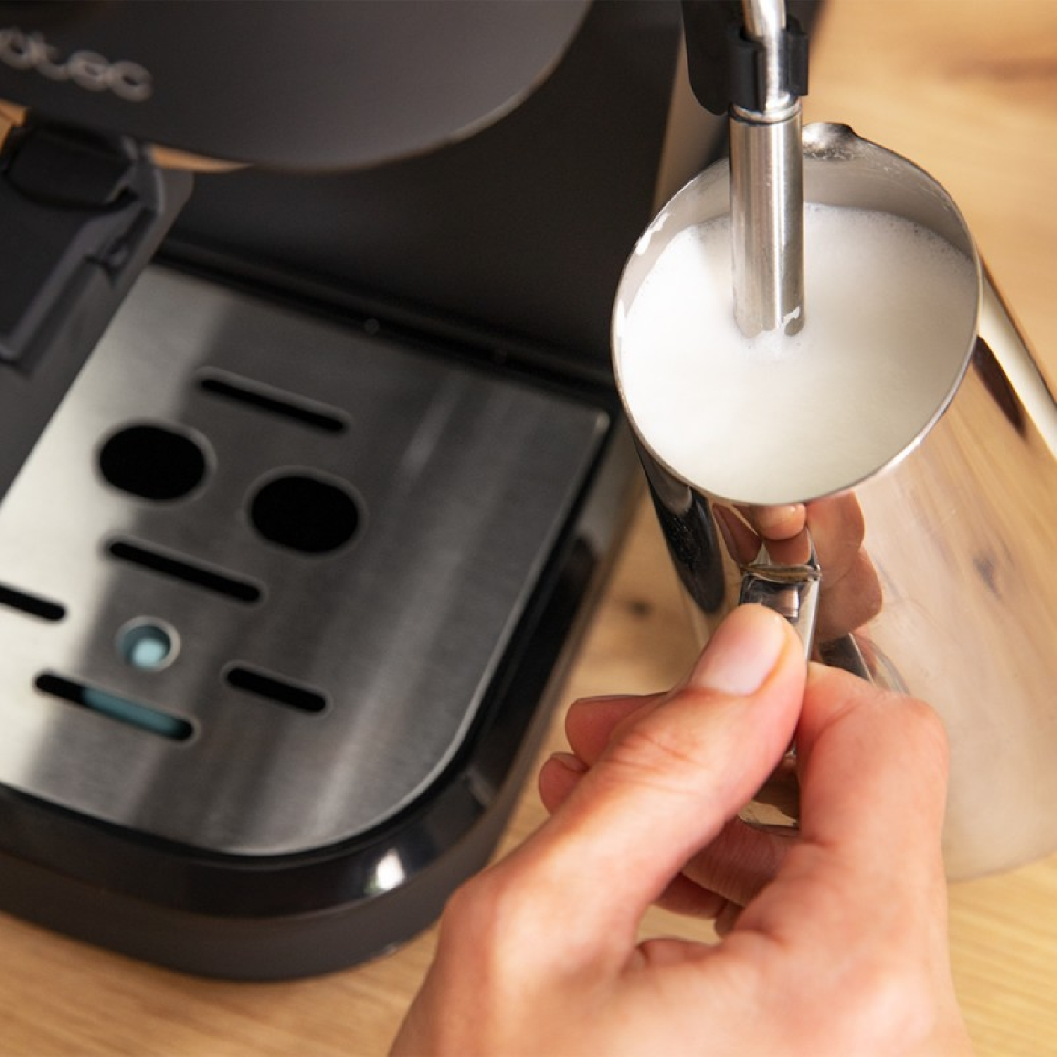 Ręka trzyma dzbanek do spieniania mleka ze stali nierdzewnej, spieniając mleko. Ekspres do kawy w tle.