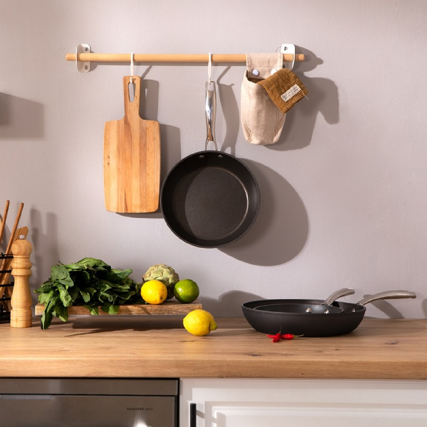 Keuken met houten snijplank, pan, koekenpan, citroenen, limoenen en een bos groen.