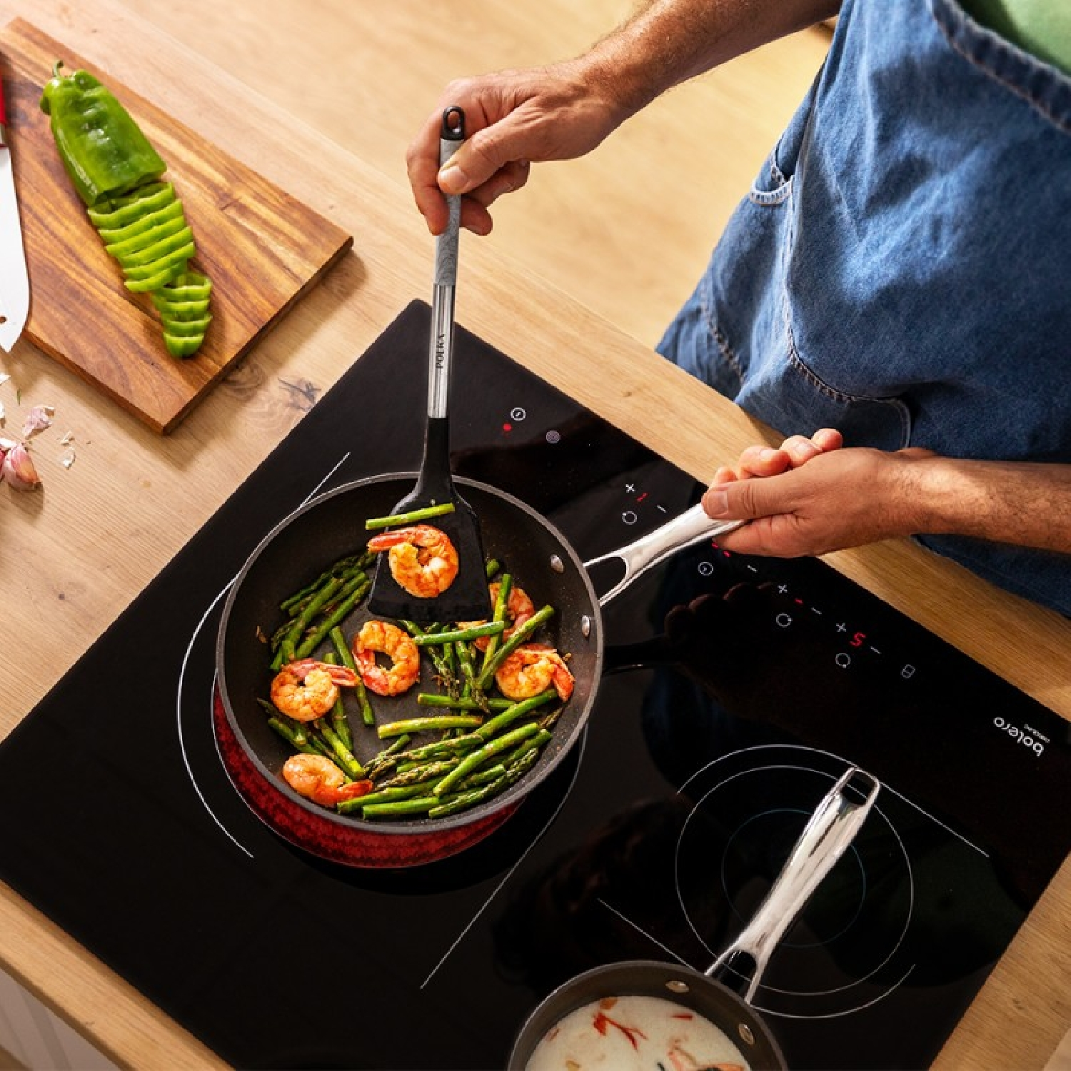 Osoba gotuje krewetki i szparagi na patelni na czarnej płycie. Deska do krojenia z papryką.