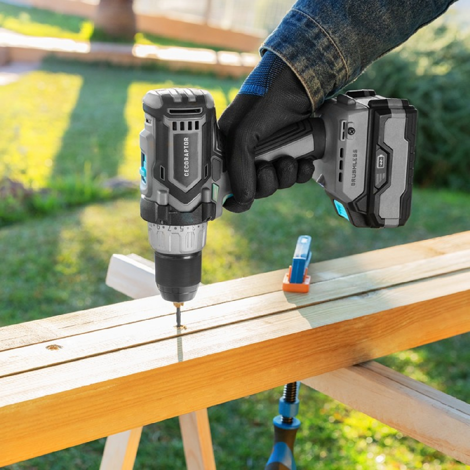 Persona che usa un trapano a batteria sul legno. Guanti neri. Tavolo di legno, erba verde.