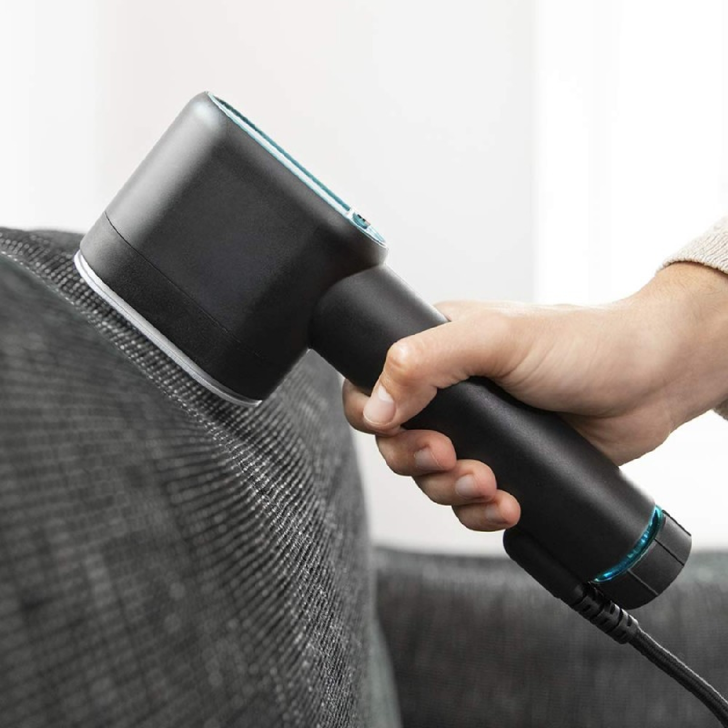 Eine Hand benutzt einen schwarzen Stoffrasierer auf einem grauen Sofa.