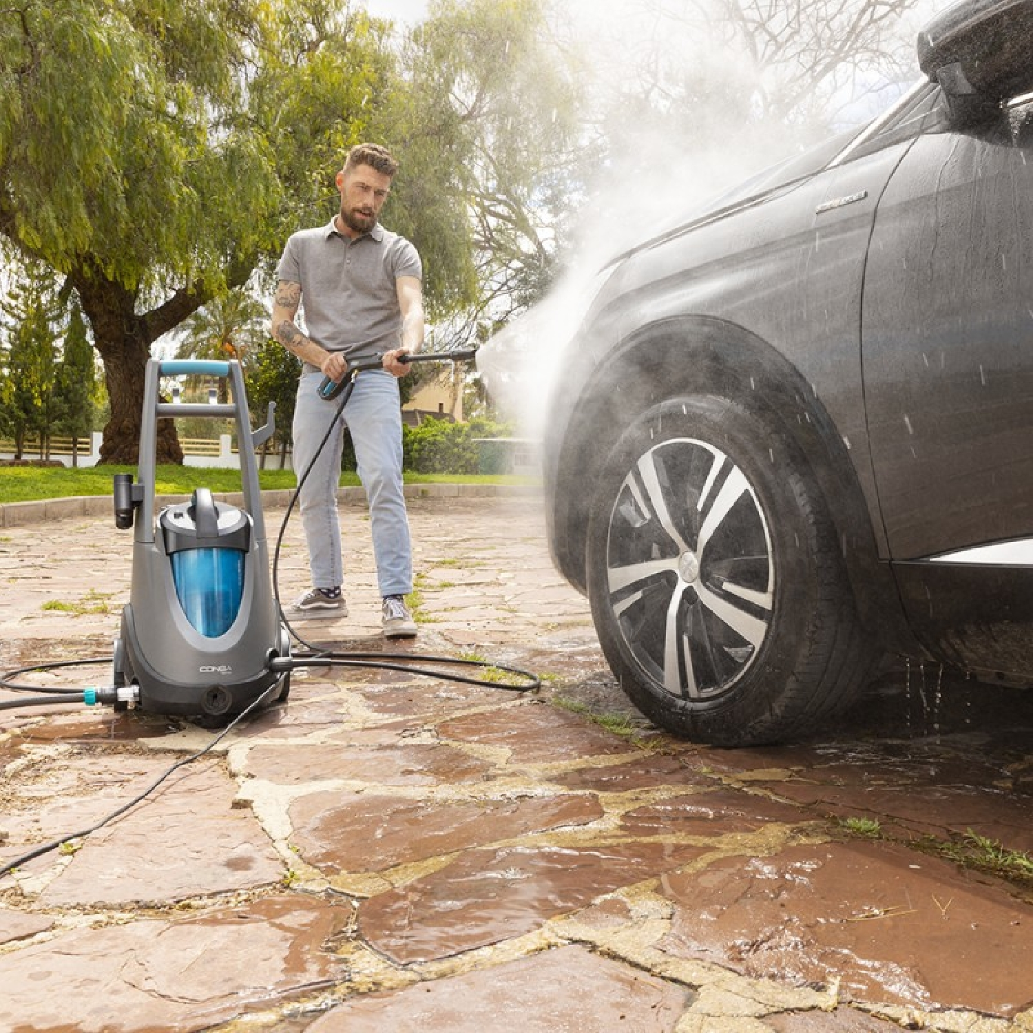 Een man maakt een donkergrijze auto schoon met een hogedrukreiniger. Water spuit op een terras.