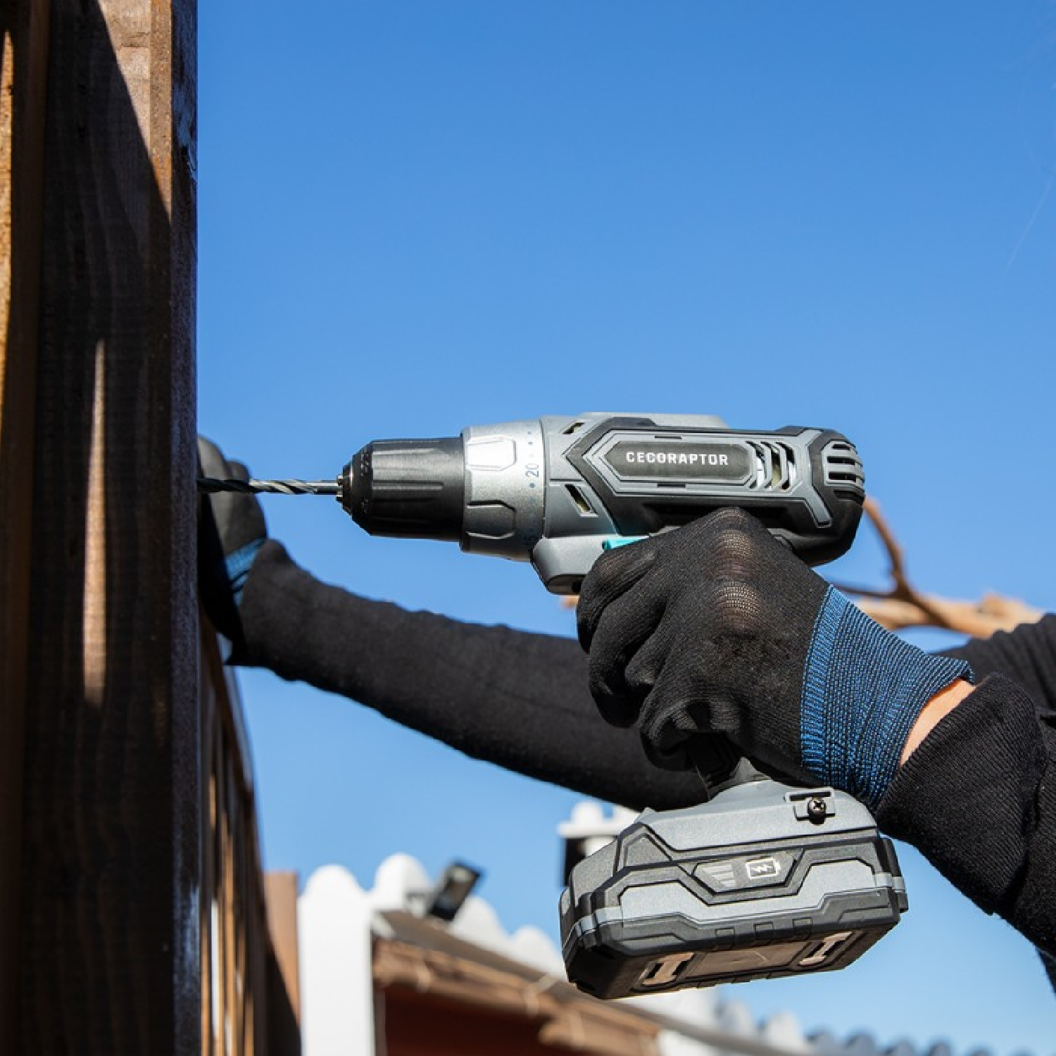 Osoba używająca wiertarki na drewnianym płocie, niebieskie niebo w tle.