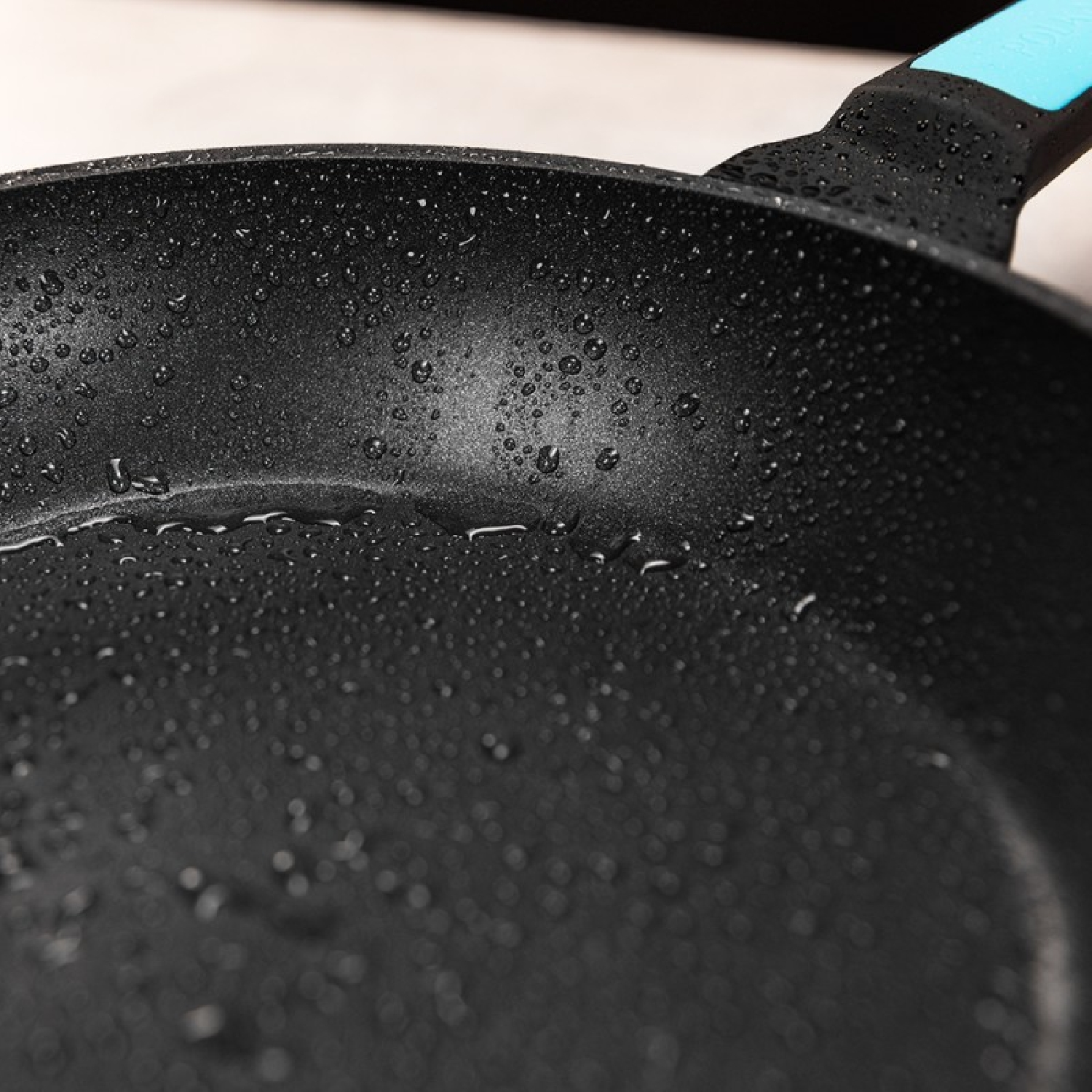 Zbliżenie patelni nieprzywierającej z kroplami wody, pokazujące jej czarną powierzchnię i niebieski uchwyt.