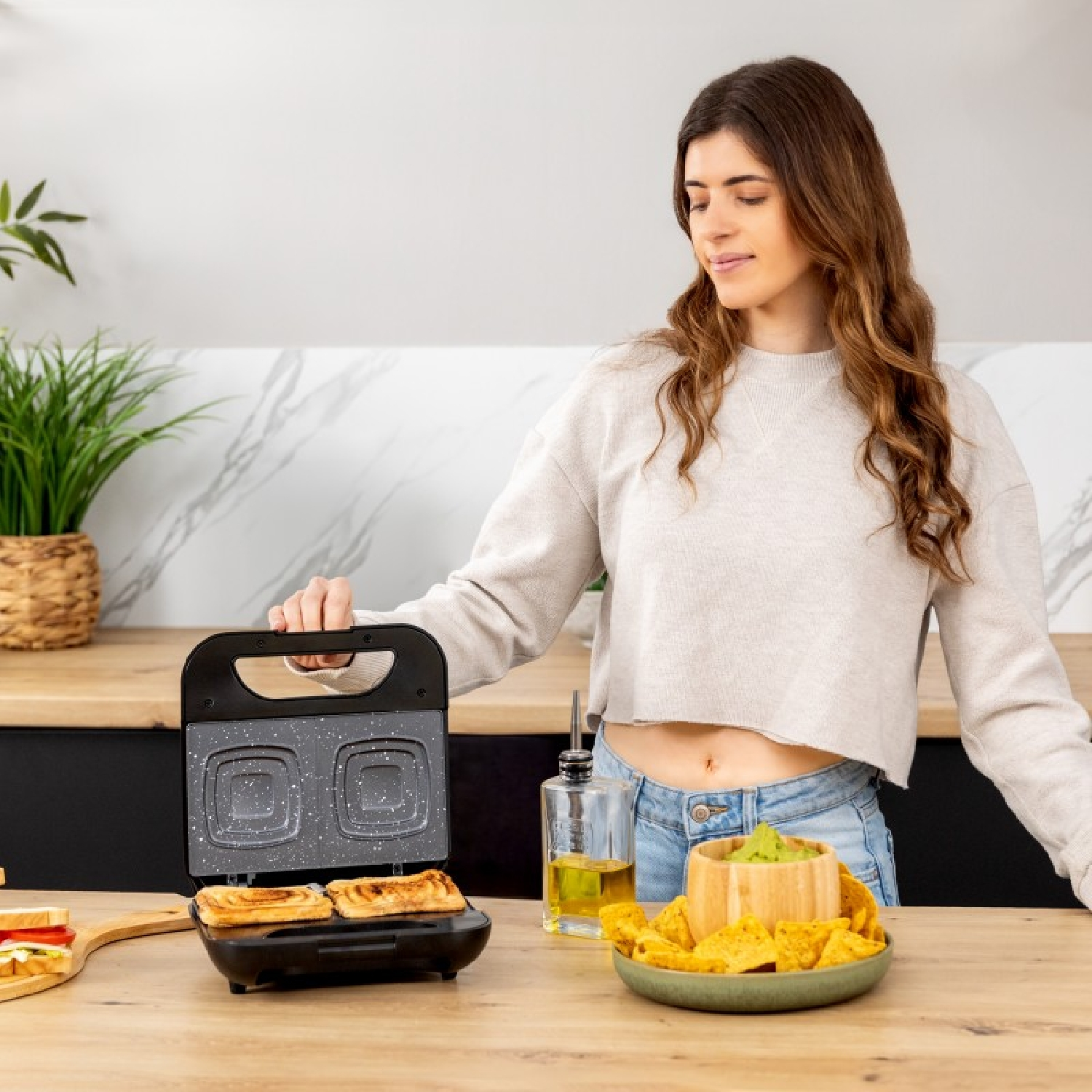 Een vrouw bekijkt een sandwichmaker met geroosterde sandwiches. Er is een kom met nacho's en olie.