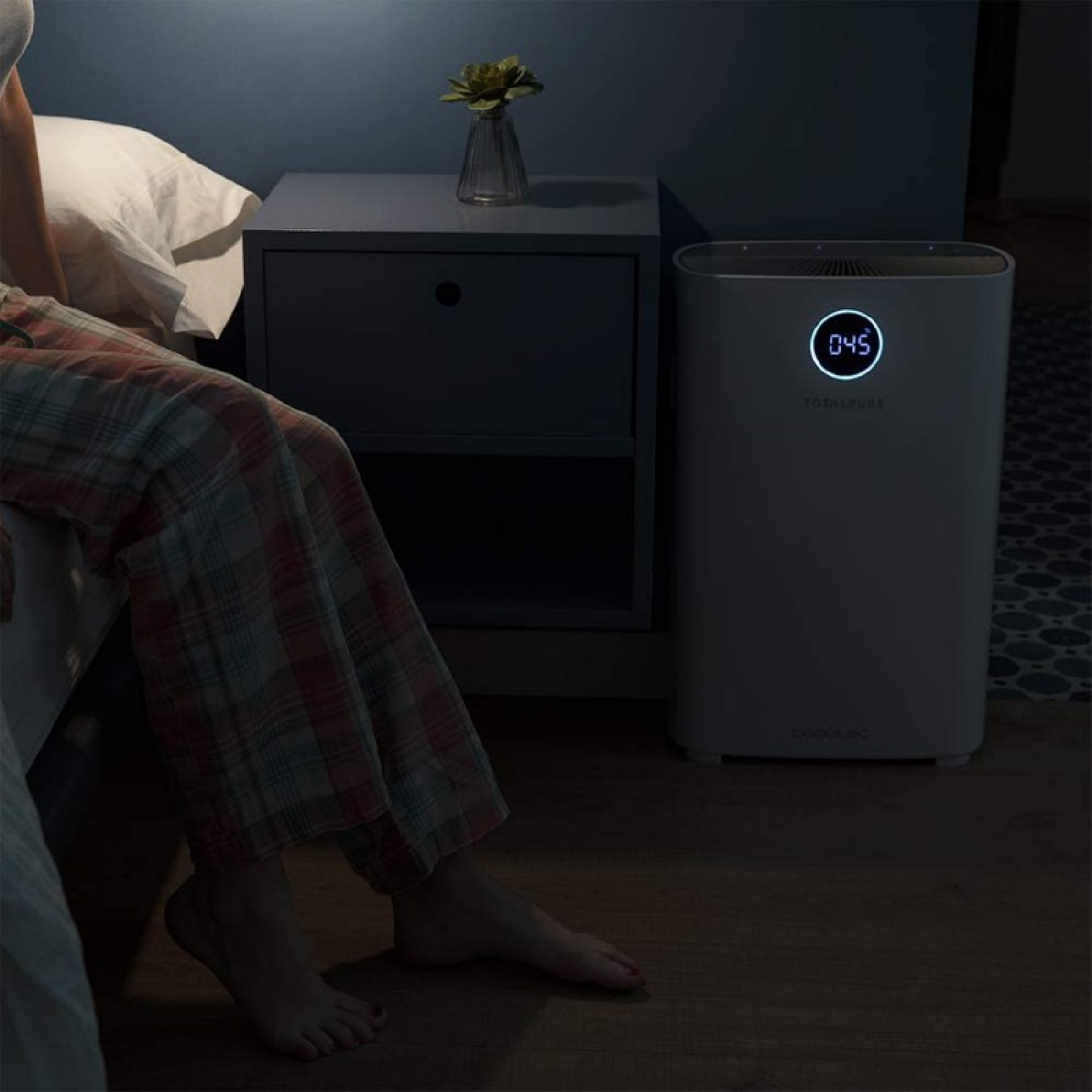 Una persona è seduta su un letto, un purificatore d'aria è accanto al letto. Un comodino sullo sfondo.