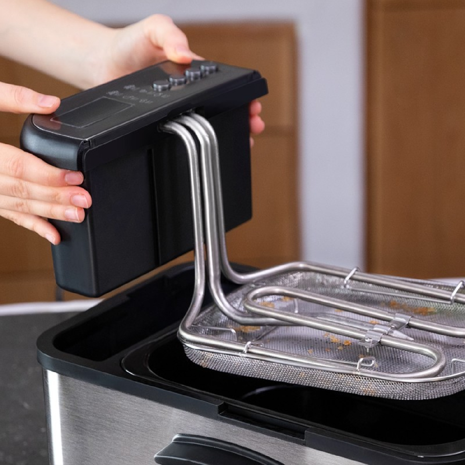 Ręce osoby dodającej czarną część do frytkownicy w kolorze stali. Drewniana szafka w tle.