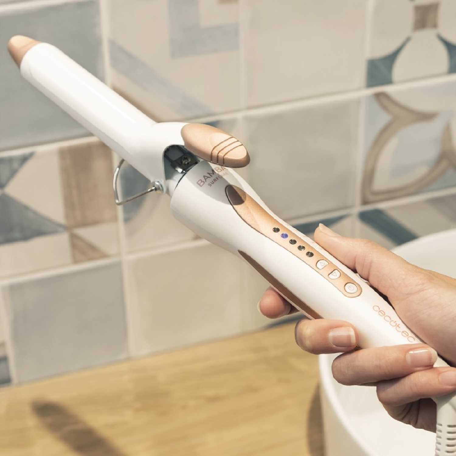 Dłoń kobiety trzyma białą i złotą lokówkę. Tło: kafelkowa ściana.