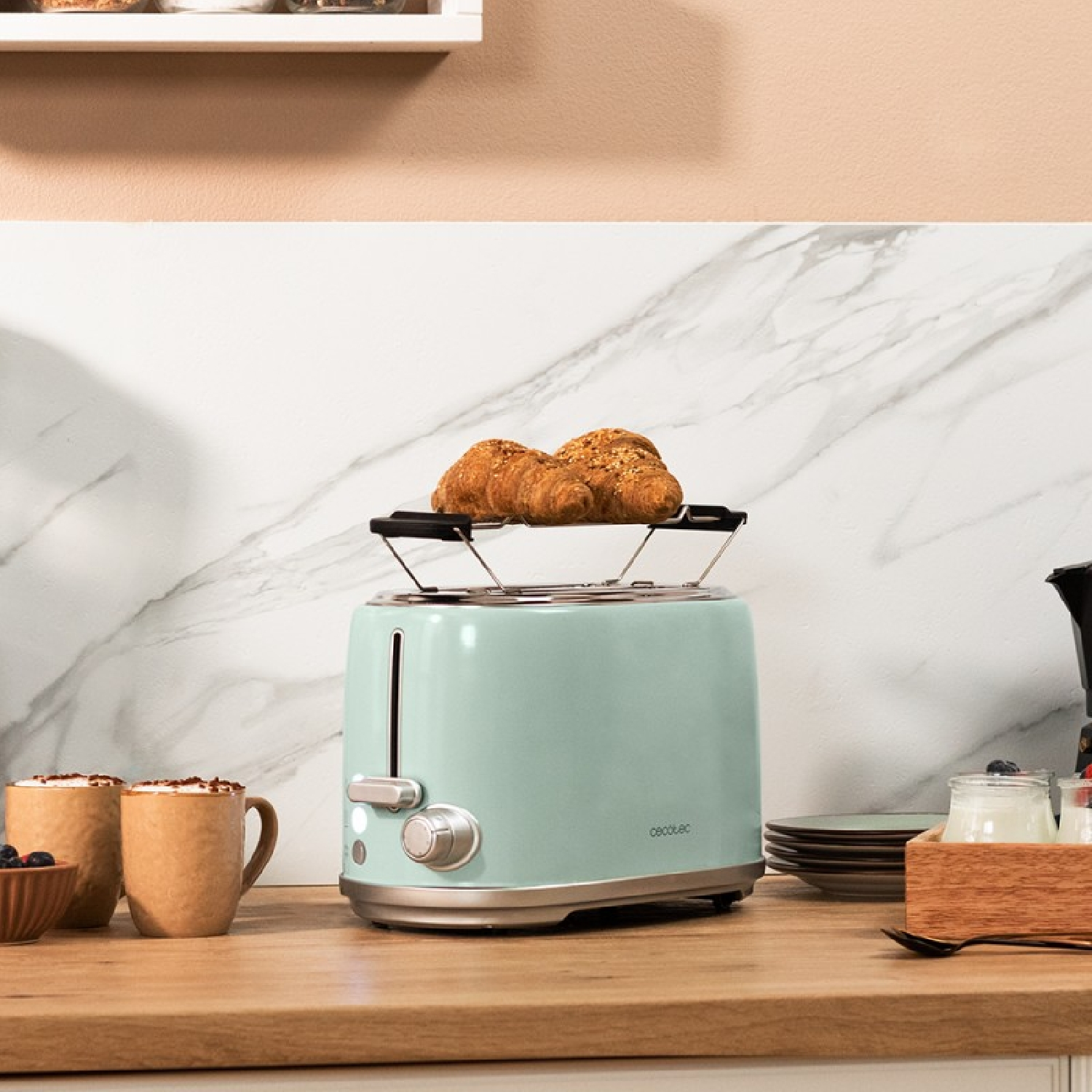 Mint green toaster with croissants on top, mugs, and other breakfast items on a counter.