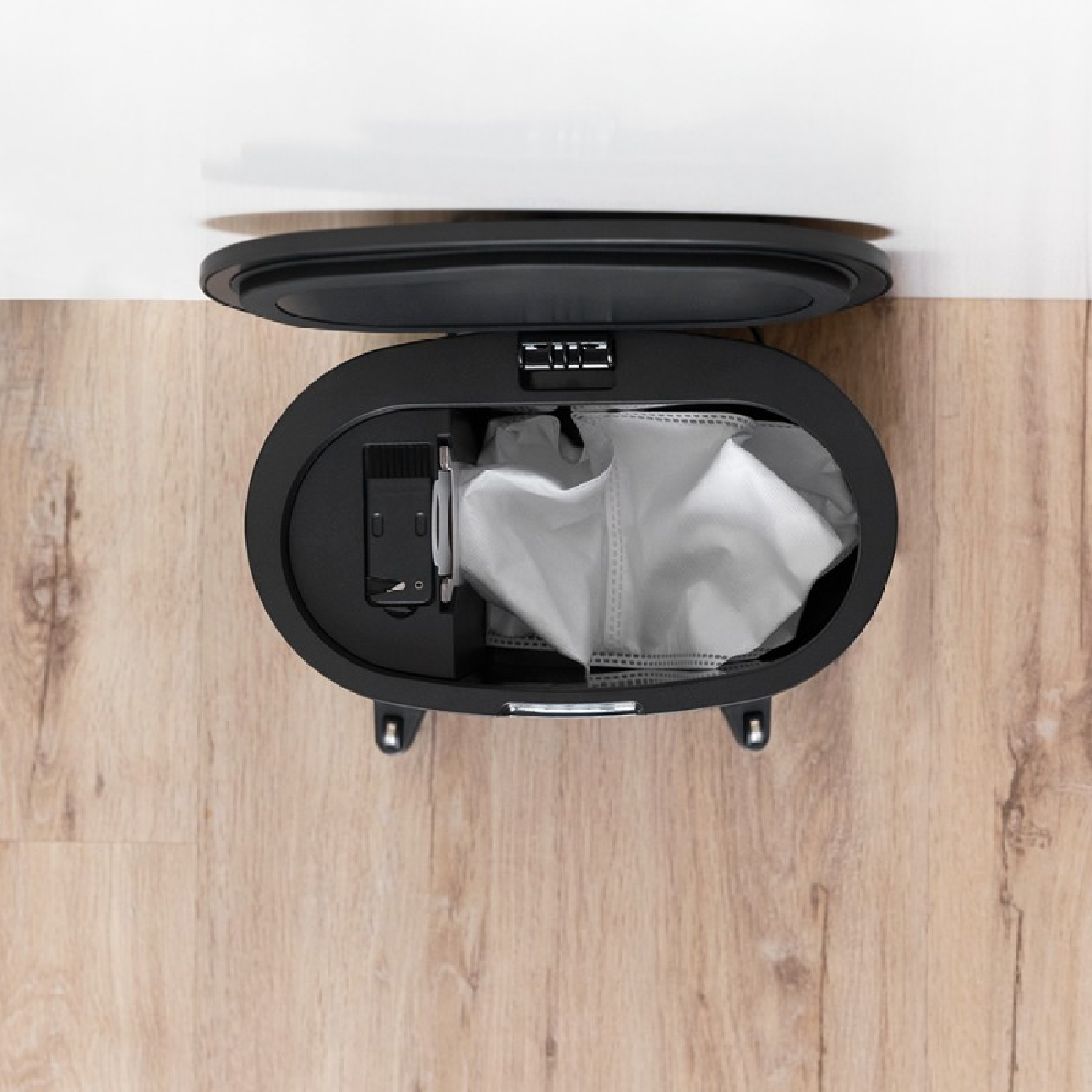 Bidone della spazzatura nero con coperchio aperto che mostra un sacco bianco all'interno, su pavimento in legno.