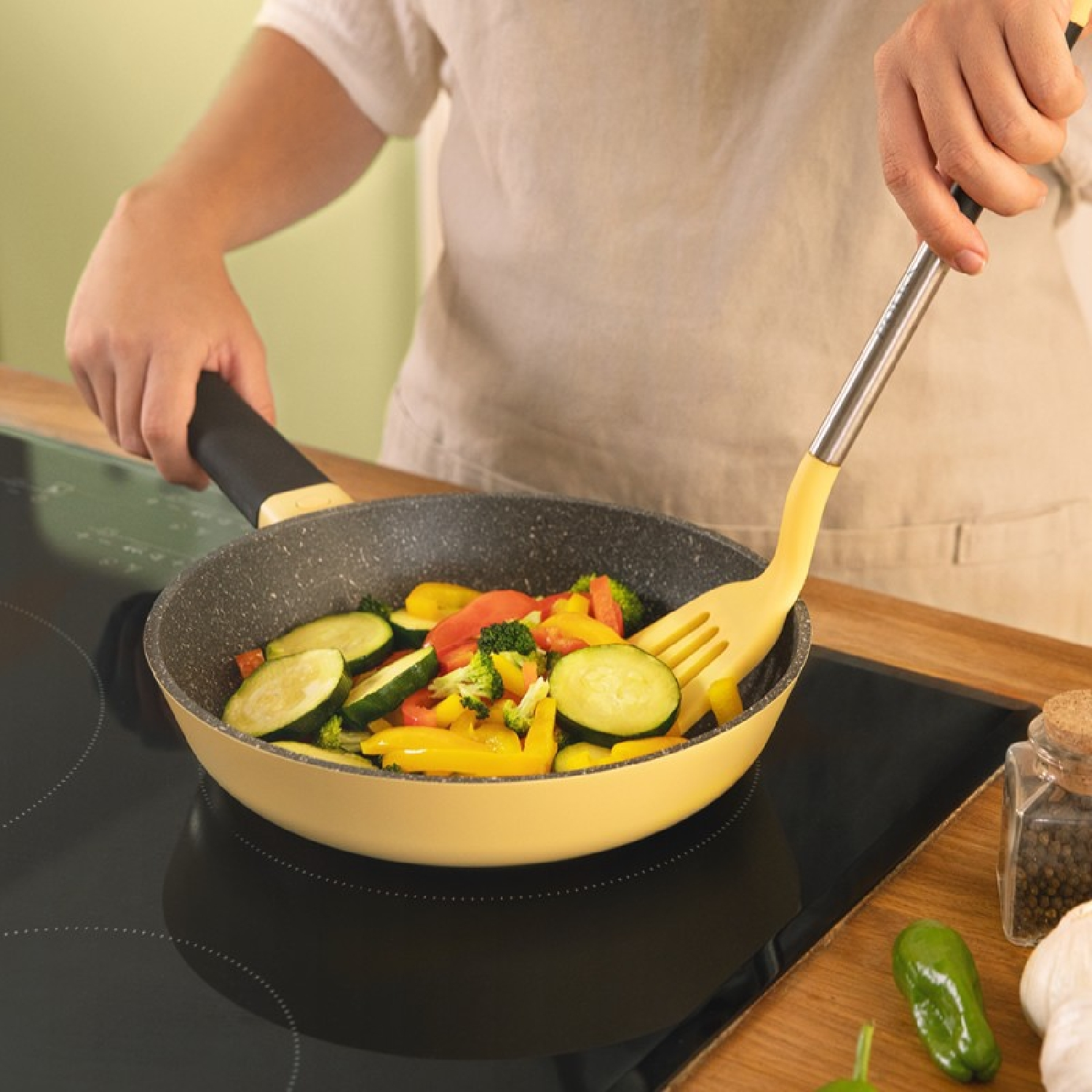 Una persona che cucina verdure in una padella gialla su un piano cottura nero.