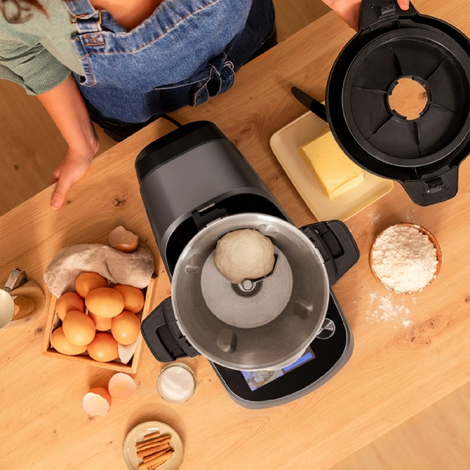 Una persona prepara masa en un electrodoméstico de cocina. Huevos, mantequilla y harina sobre la mesa.