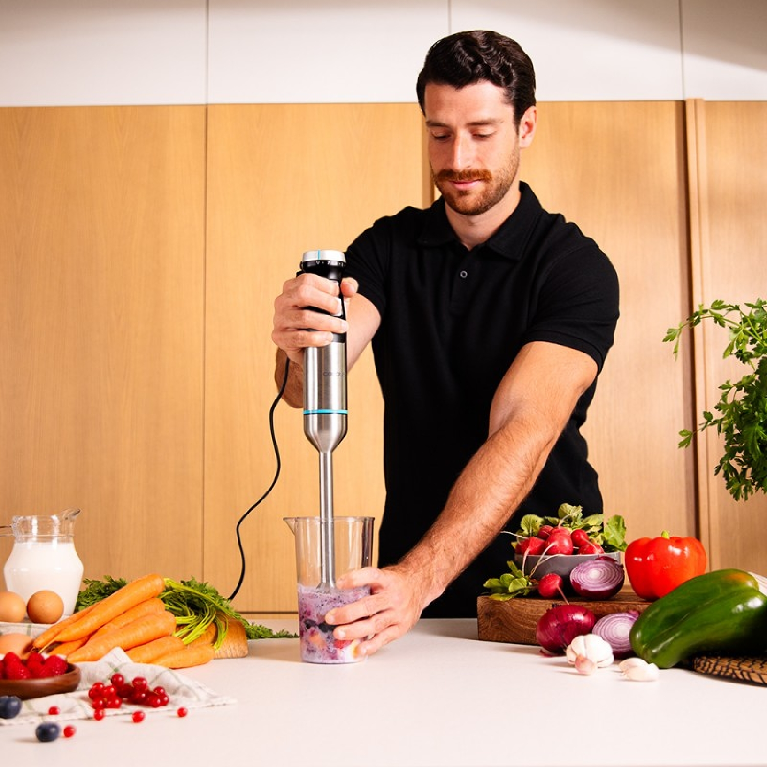 Uomo che frulla ingredienti in un bicchiere con un frullatore a immersione. Verdure e frutta sul tavolo.