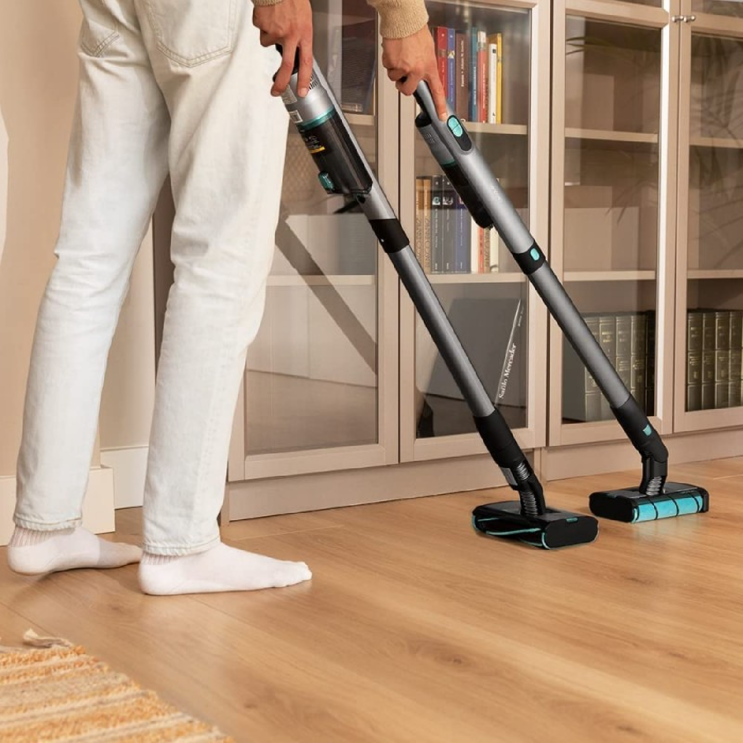 A man is cleaning a wooden floor with two cordless vacuum cleaners. The vacuums are grey and black.