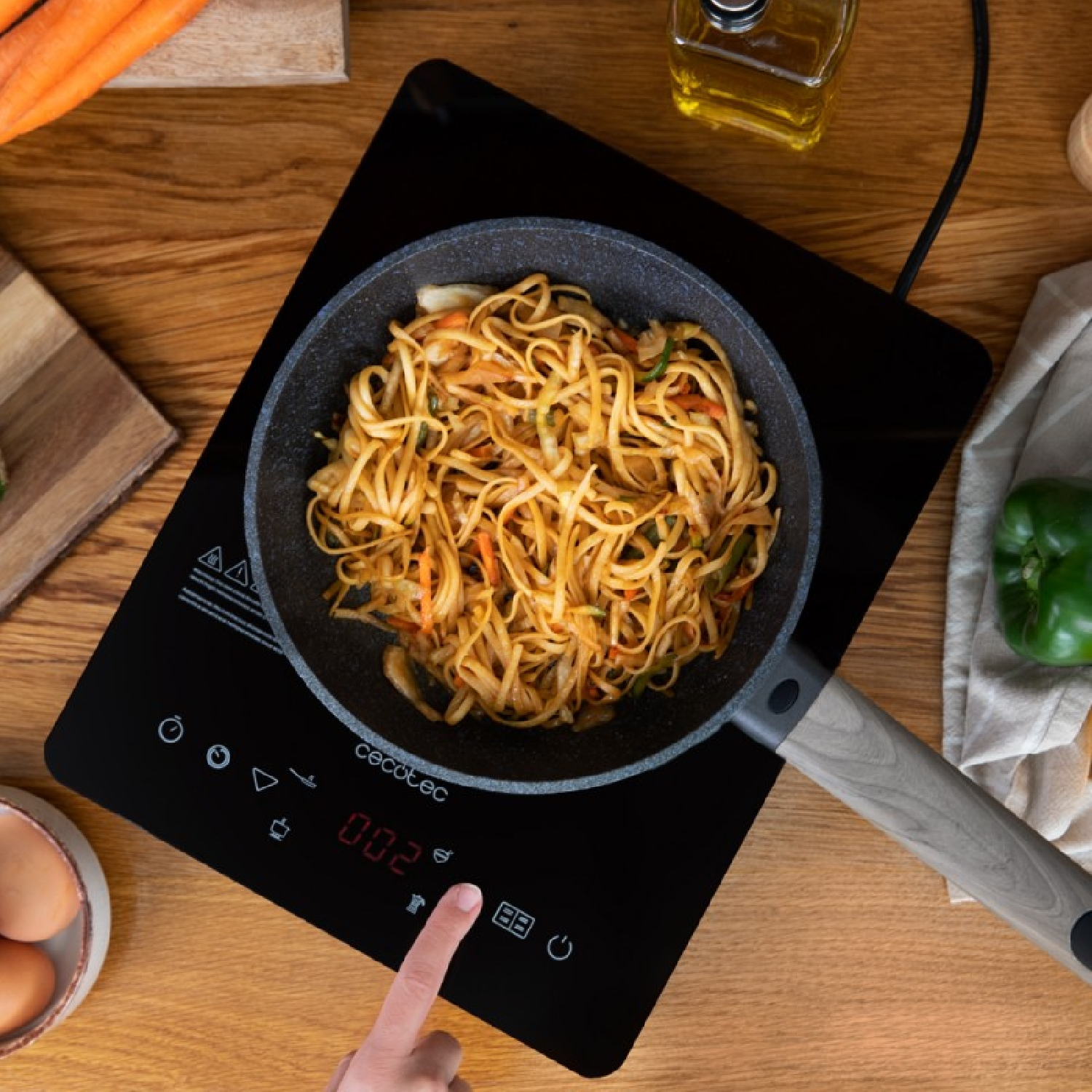 Ręka używa czarnej płyty indukcyjnej. Na płycie patelnia z makaronem. Warzywa z boku.