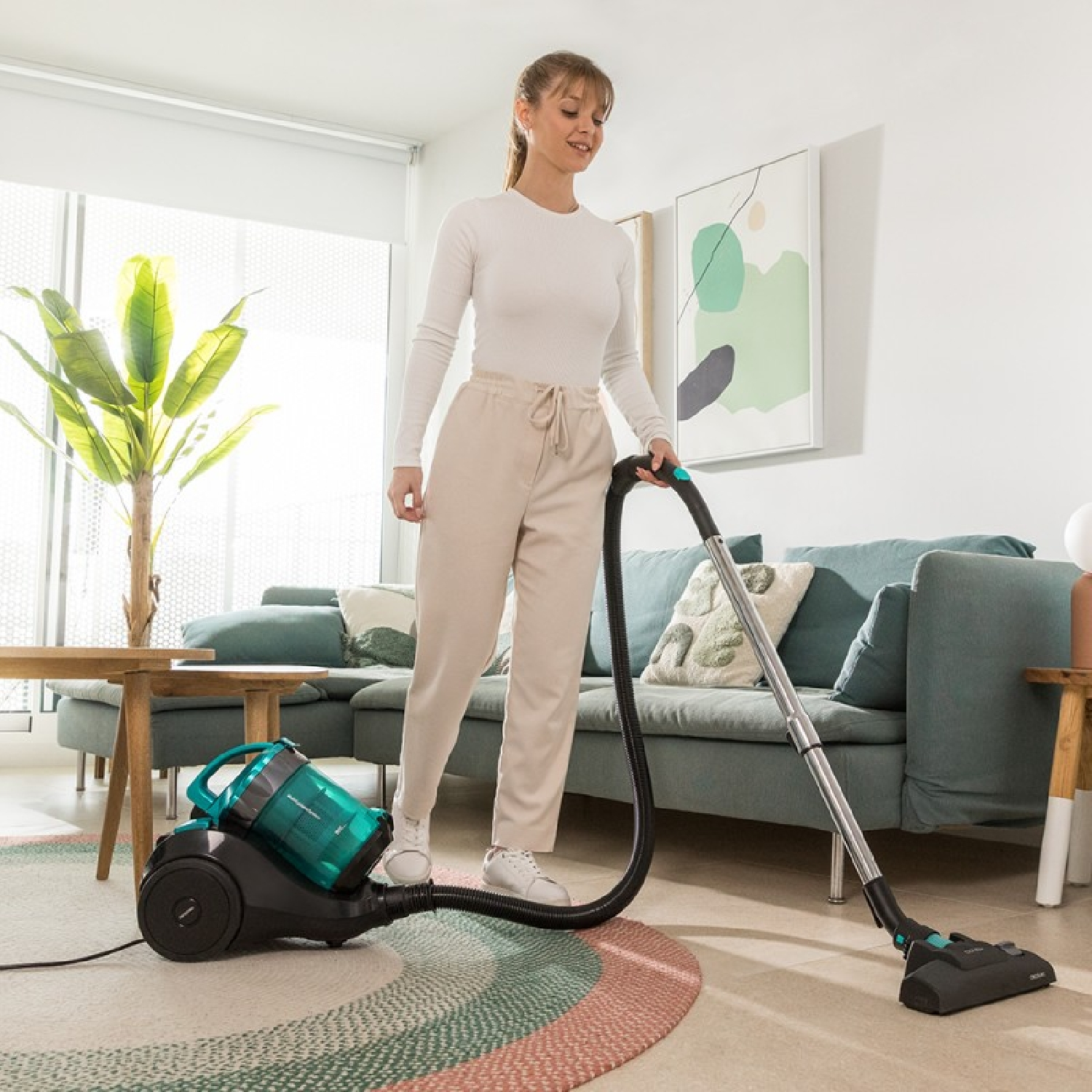 Frau saugt ein Zimmer. Staubsauger auf dem Boden, Frau hält Schlauch, Sofa im Hintergrund.
