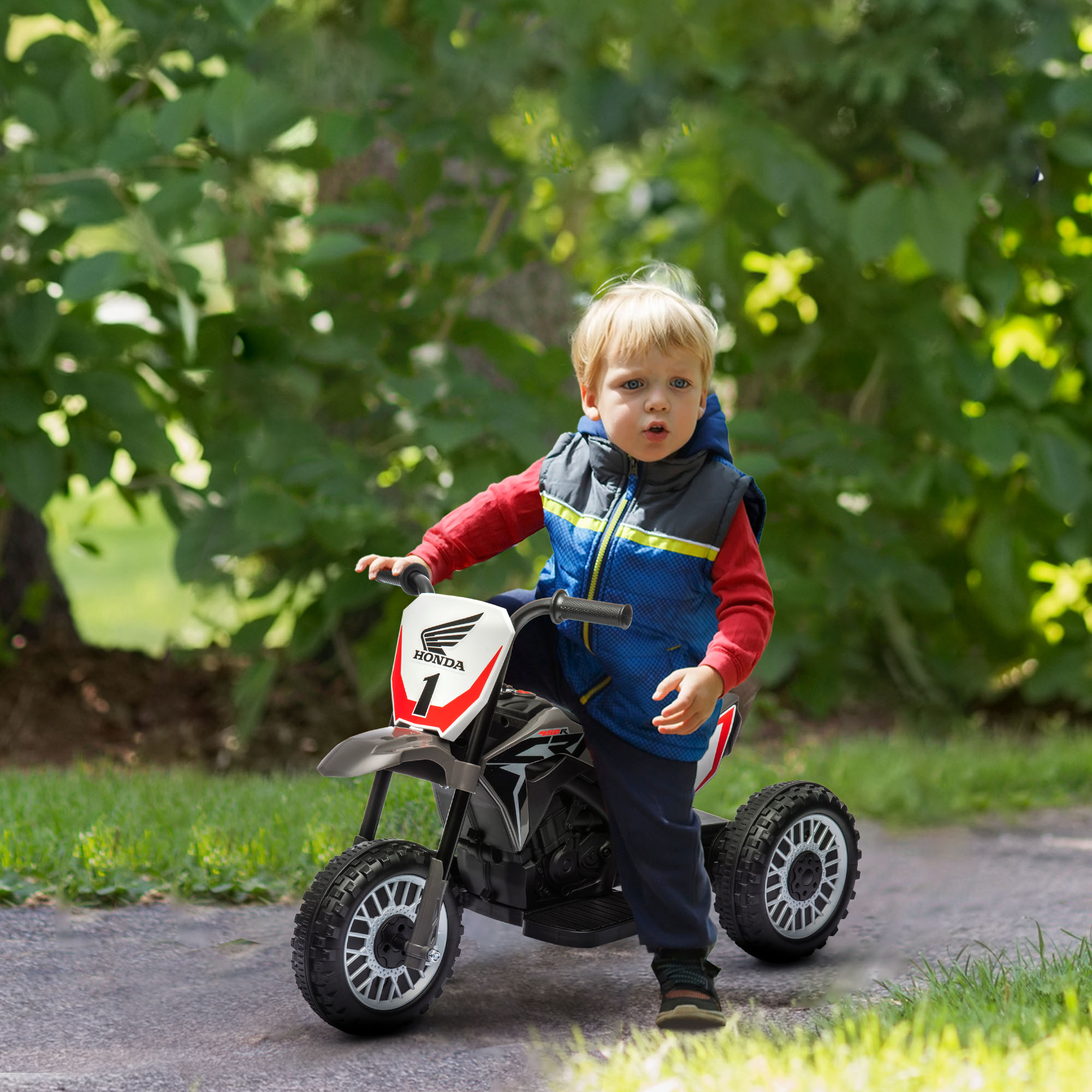 Un niño pequeño montando una moto de juguete en un camino pavimentado. Árboles y hierba verde en el fondo.