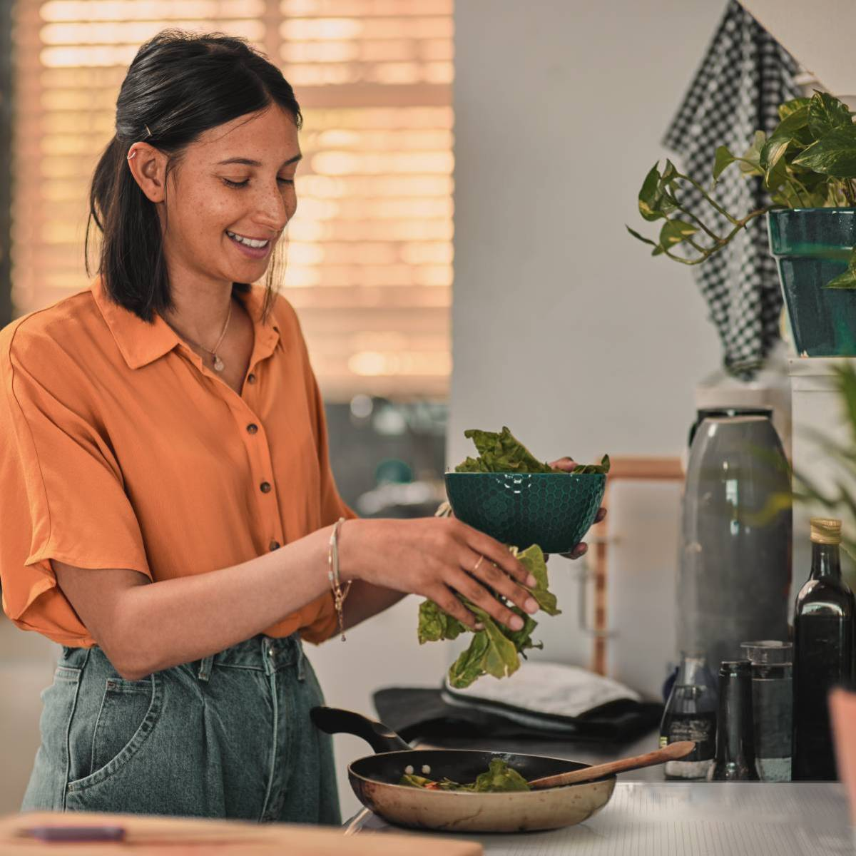 Una mujer con una camisa naranja prepara una ensalada en la cocina. Una sartén está en la estufa. Verduras al fondo.