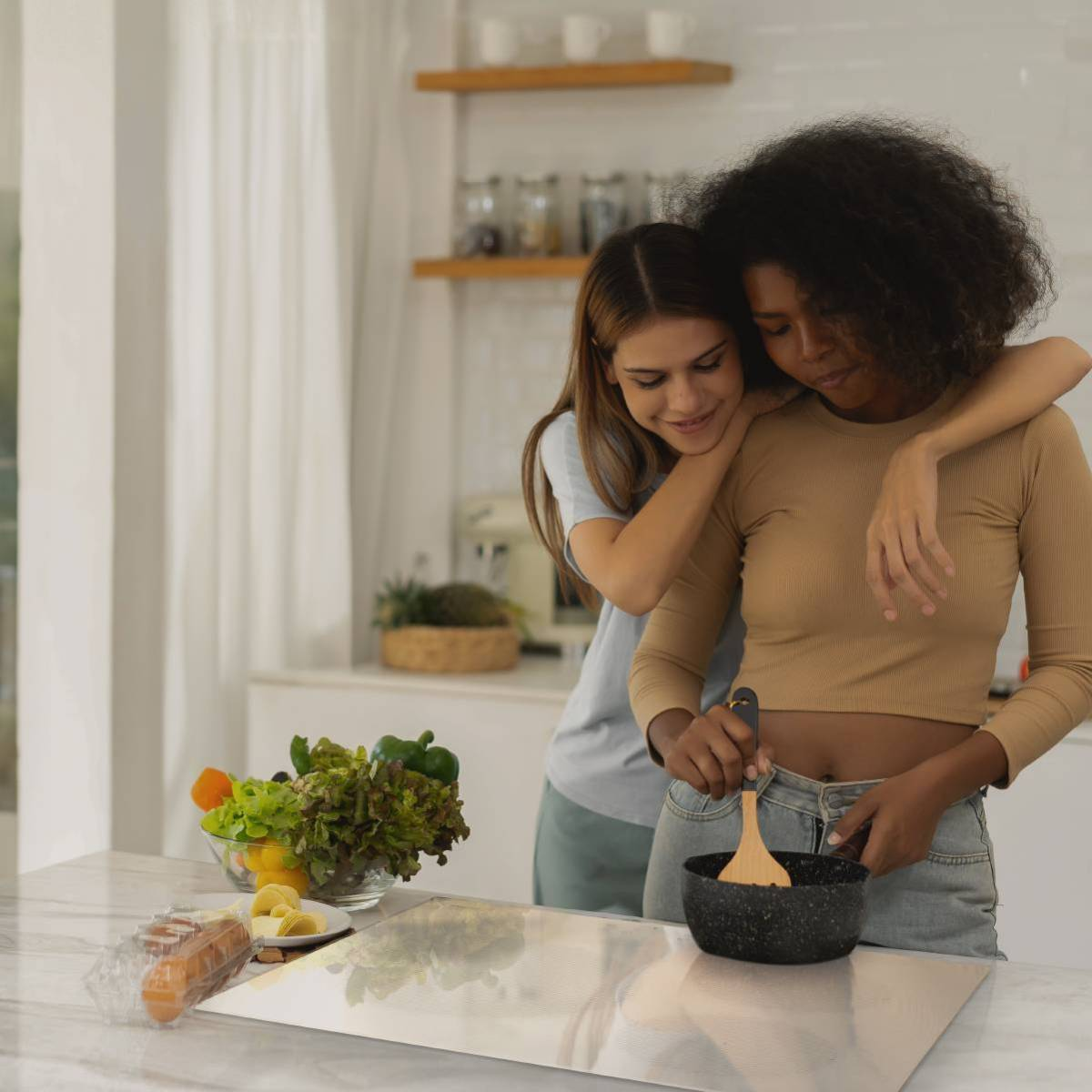 Dos mujeres cocinando en una cocina, una removiendo una olla, la otra abrazándola, con ingredientes cerca.