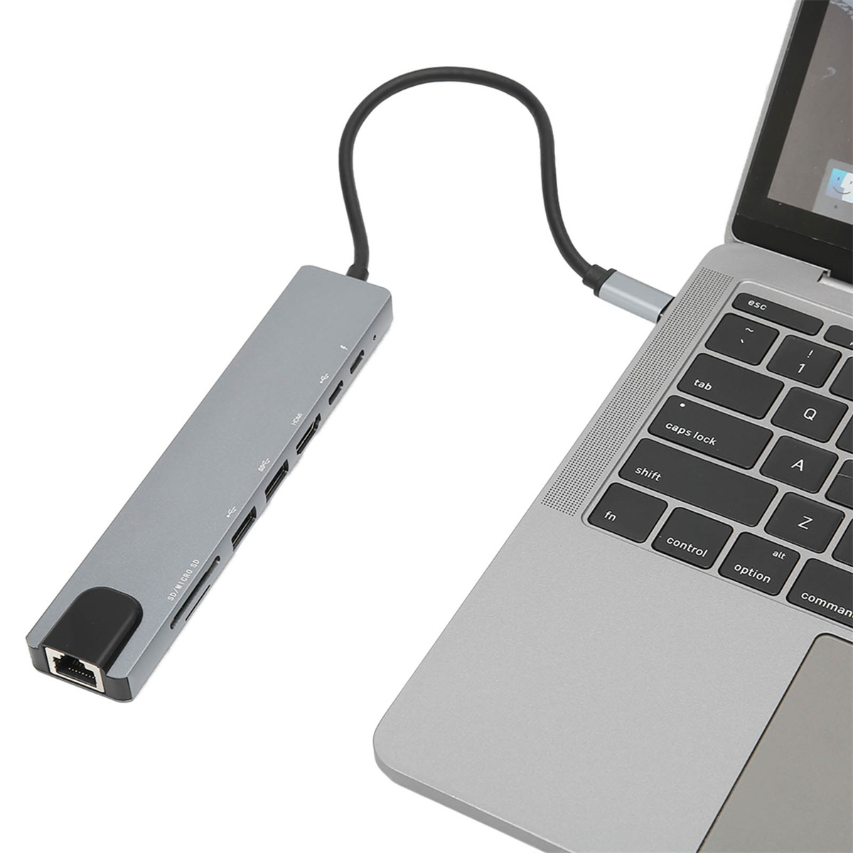 Ein grauer USB-C-Hub, der mit einem Laptop verbunden ist. Mehrere Anschlüsse sichtbar. Schwarzes Kabel.