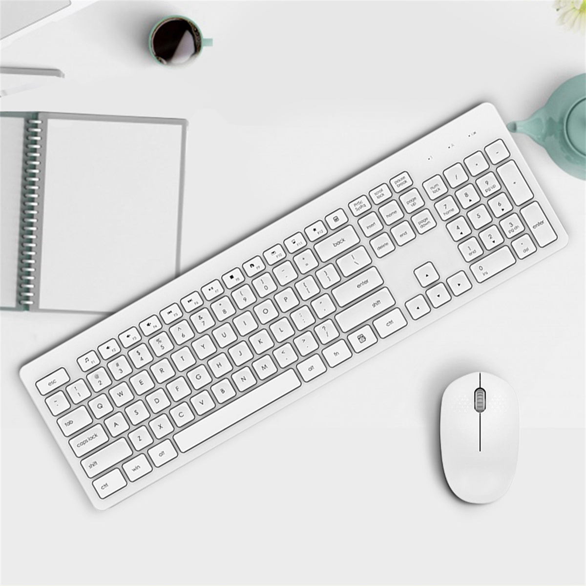 Weiße kabellose Tastatur und Maus auf weißer Oberfläche. Notizblock und Tasse sichtbar.
