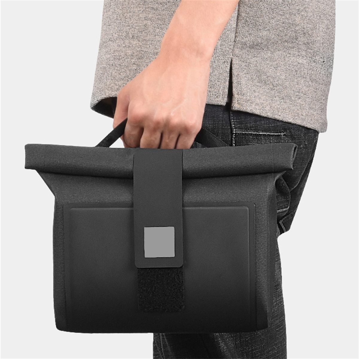 Eine schwarze Umhängetasche mit Klappe und Griff. Eine Person hält die Tasche.