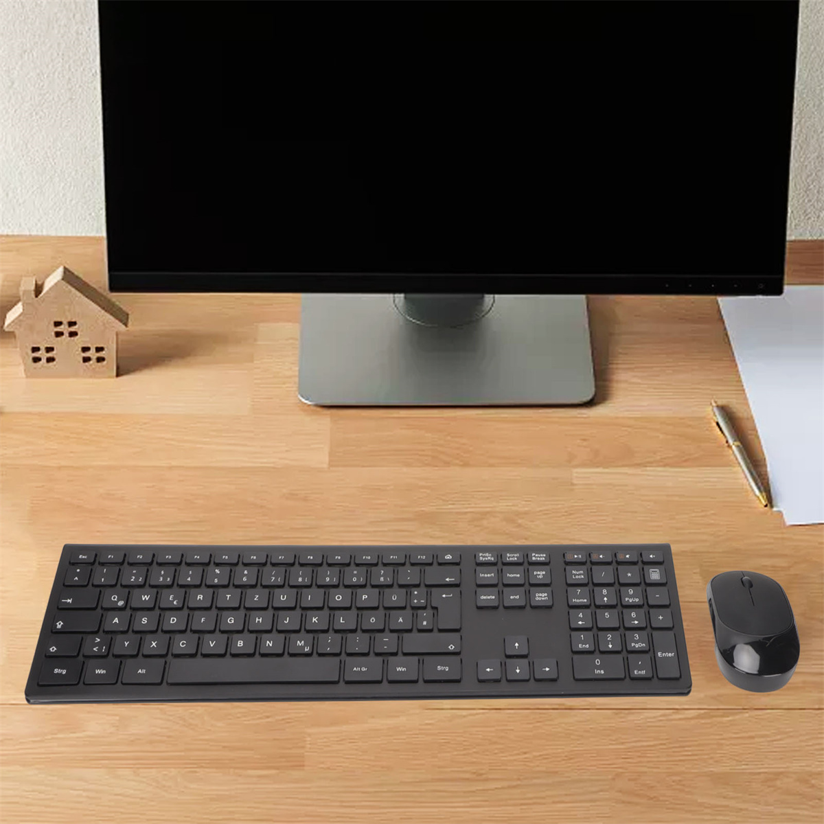 Ein Schreibtisch-Setup mit schwarzer Tastatur, Maus, Monitor und einer kleinen Holzhaus-Dekoration auf einem Holztisch.