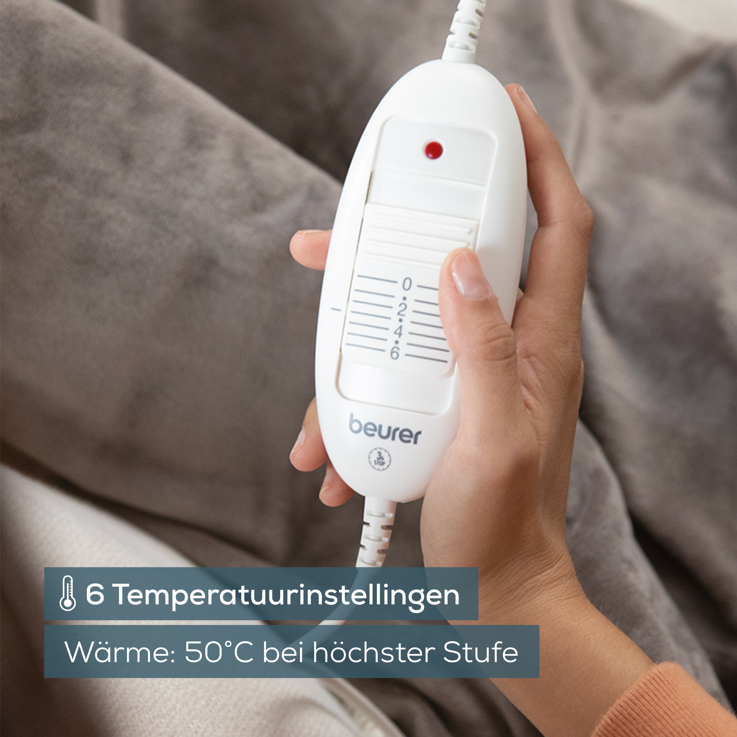 A hand holding a white electric blanket controller on a gray blanket.