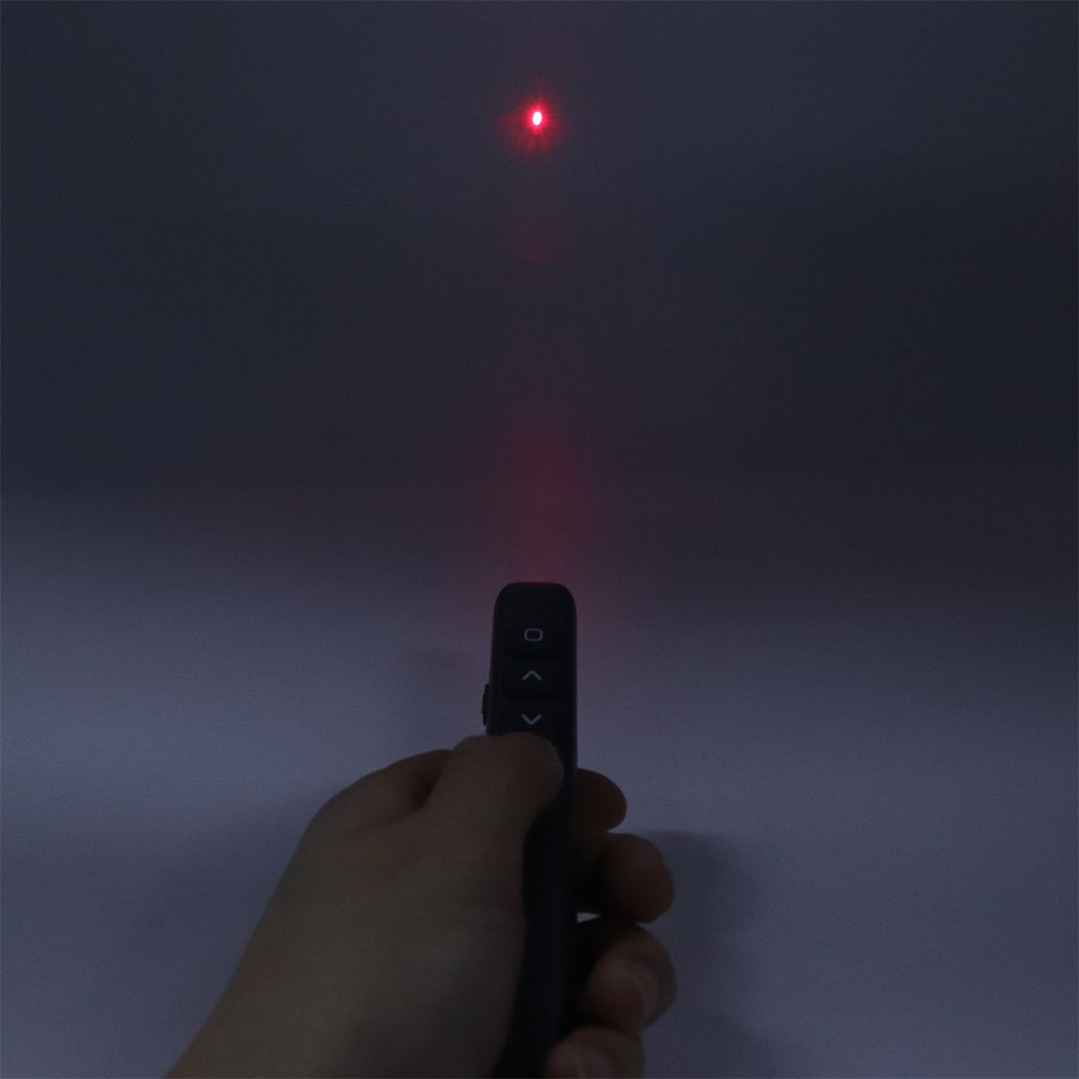 Hand hält schwarze Fernbedienung, die roten Laserpunkt zeigt. Dunkler Hintergrund.