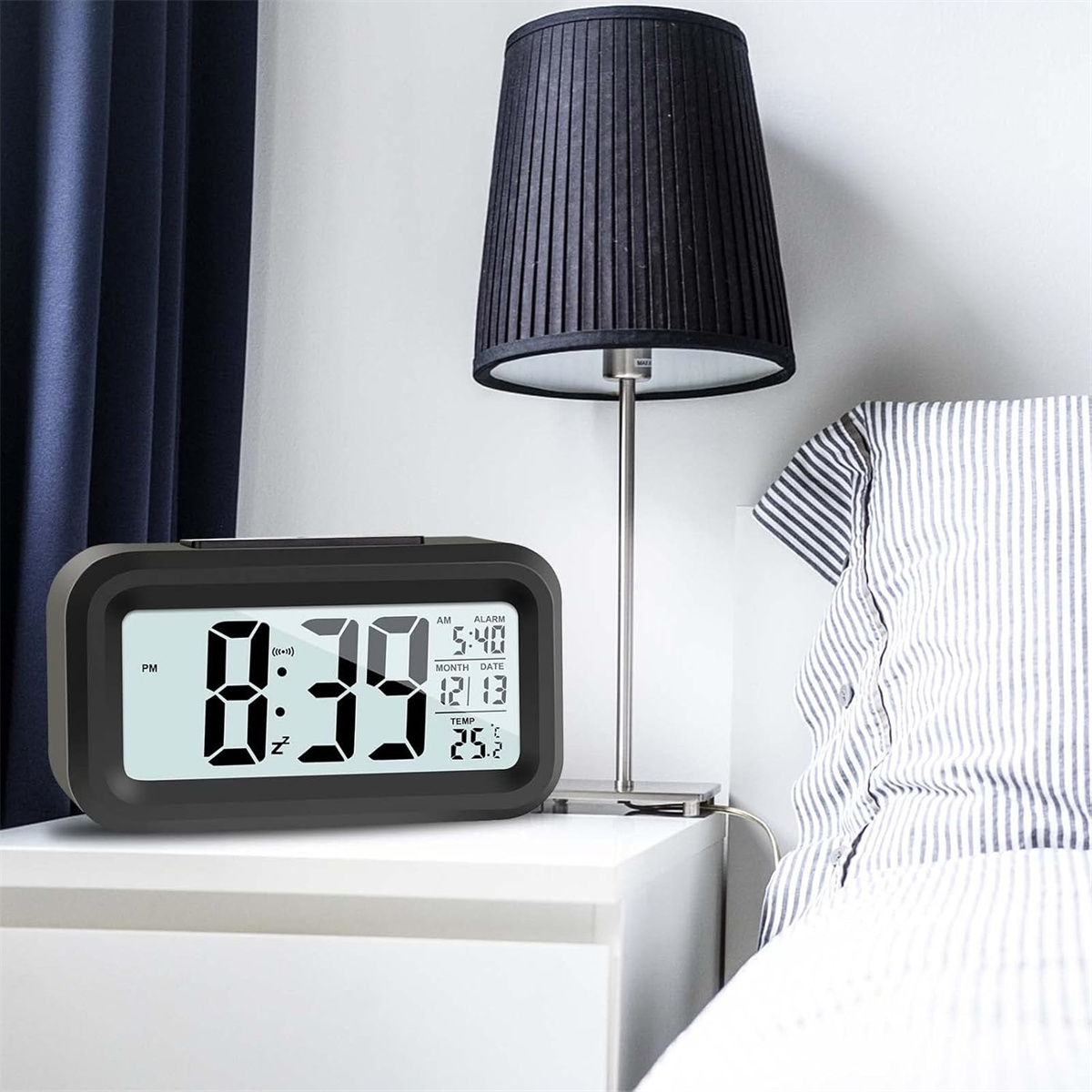 Digitaler Wecker auf einem Nachttisch, Lampe und Bett im Hintergrund.
