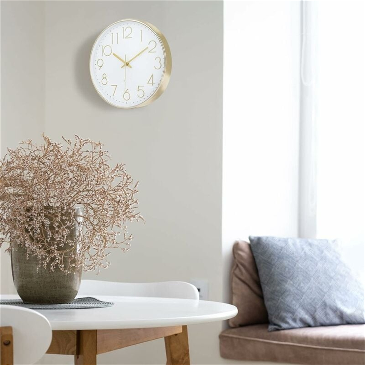 Eine goldgerahmte Uhr an einer grauen Wand über einer Vase mit getrockneten Pflanzen.