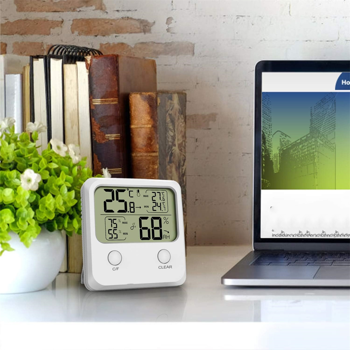 Weißes digitales Thermometer, das Temperatur und Luftfeuchtigkeit anzeigt, mit Büchern, Pflanze und Laptop in der Nähe.
