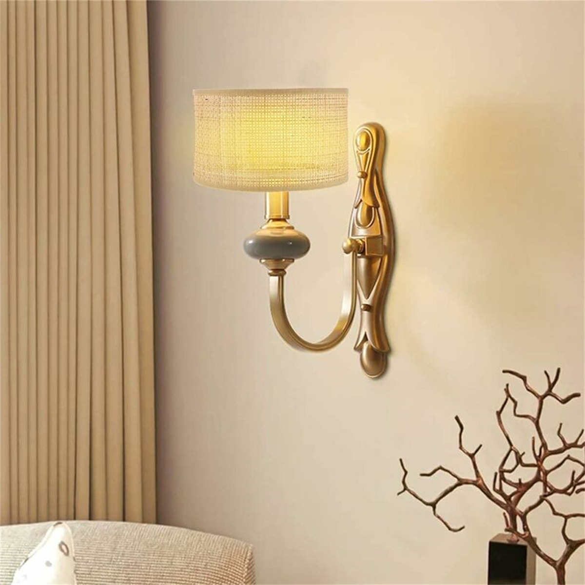 Eine Wandlampe mit beigem Lampenschirm, goldfarbener Halterung und dekorativen Zweigen daneben.