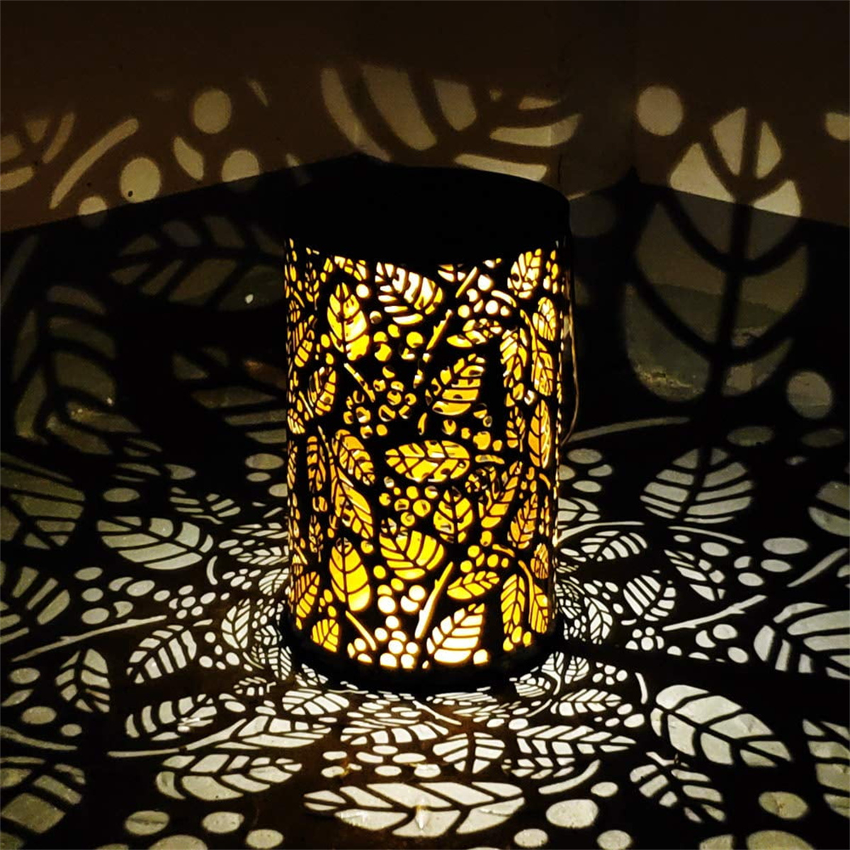 Zylindrische Lampe mit blattartigen Mustern, die Schatten werfen. Sie leuchtet und erzeugt einen warmen, einladenden Schein.