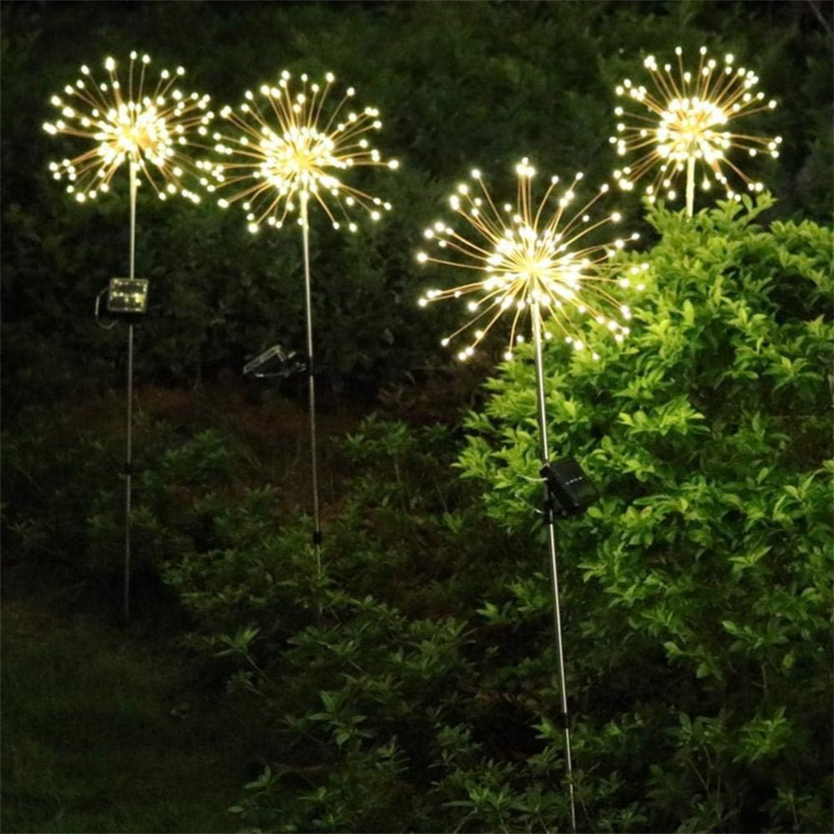 Vier dekorative Außenleuchten in Form von Feuerwerk, die einen dunkelgrünen Busch beleuchten.