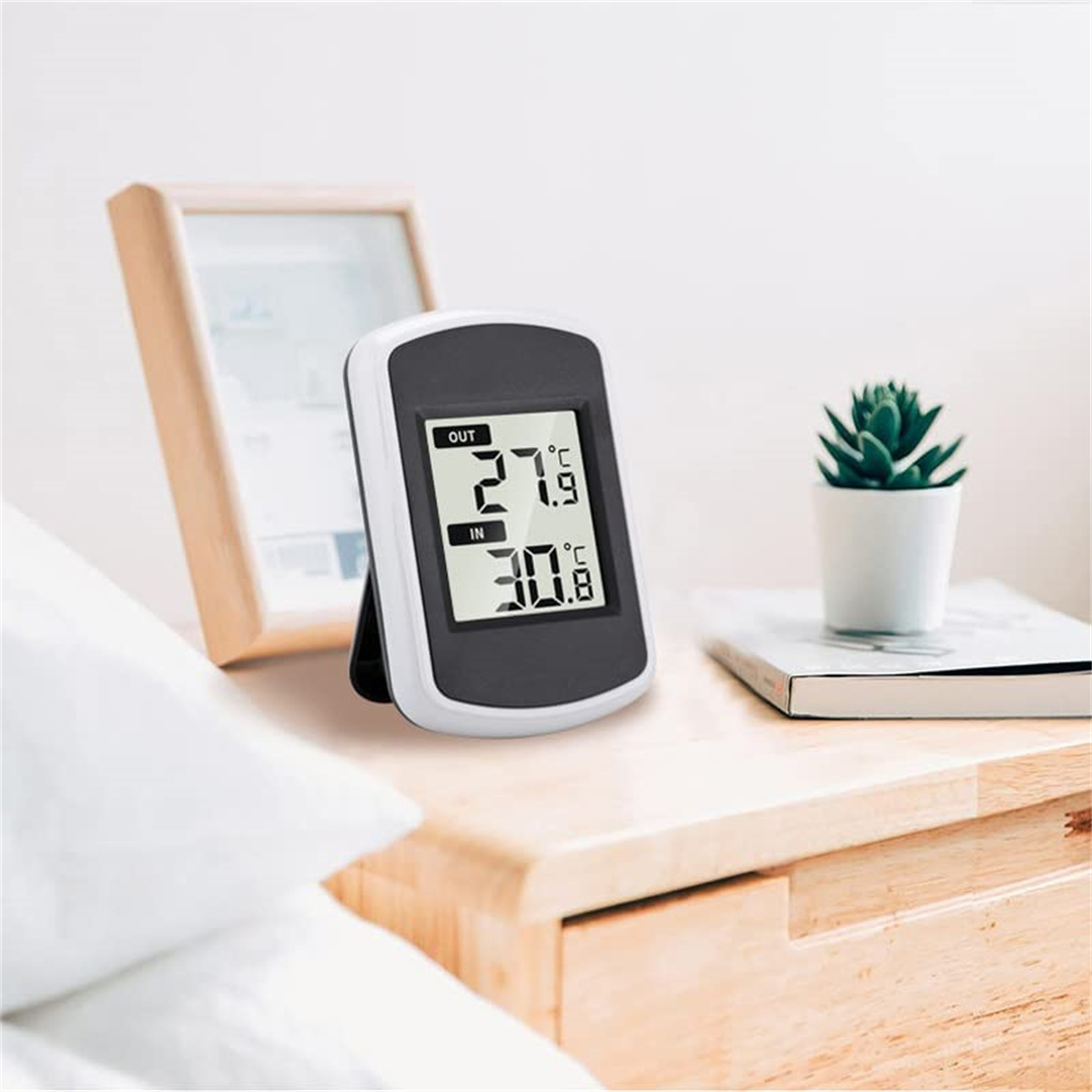 Schwarz-weißes digitales Thermometer auf einem Nachttisch, das die Temperatur anzeigt, mit einer Pflanze in der Nähe.
