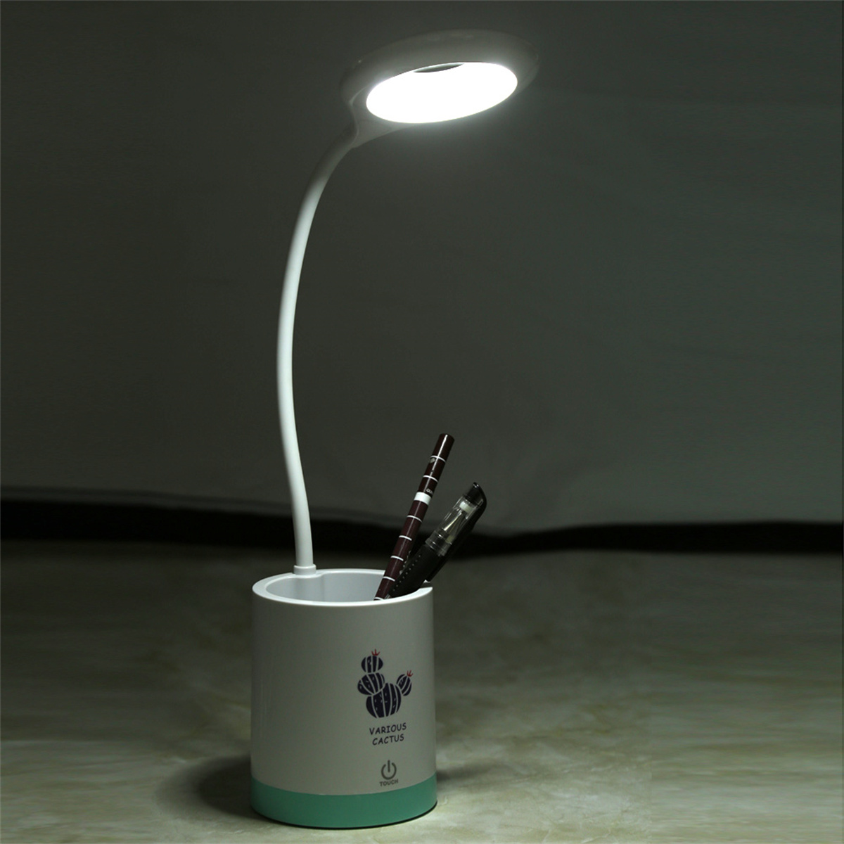 Schreibtischlampe mit Stiften in einem Halter. Die Lampe ist weiß, mit rundem Licht. Der Halter ist weiß mit grünem Sockel.
