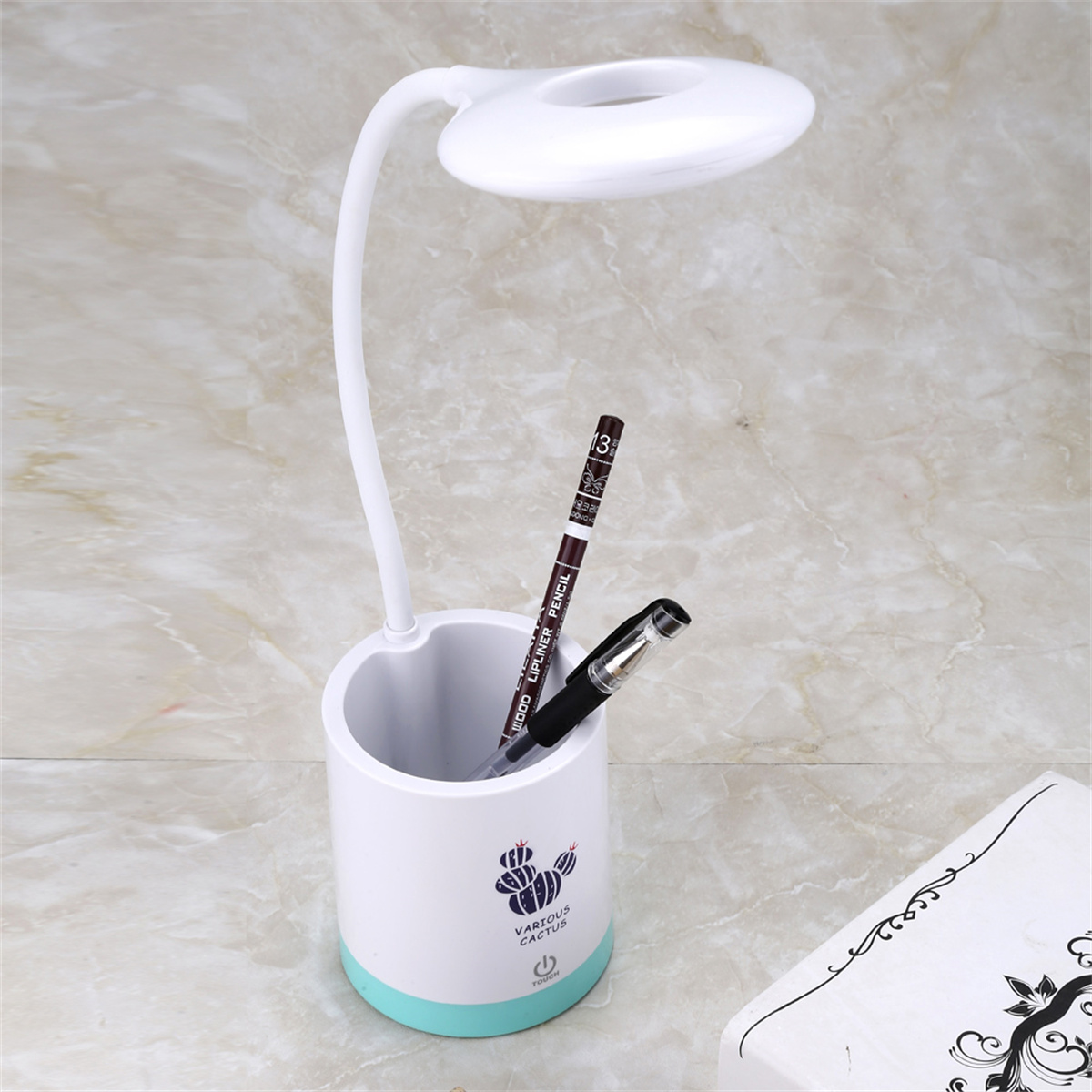 Weiße Schreibtischlampe mit Stifthalter, zwei Stiften und einem Buch auf marmorähnlicher Oberfläche.