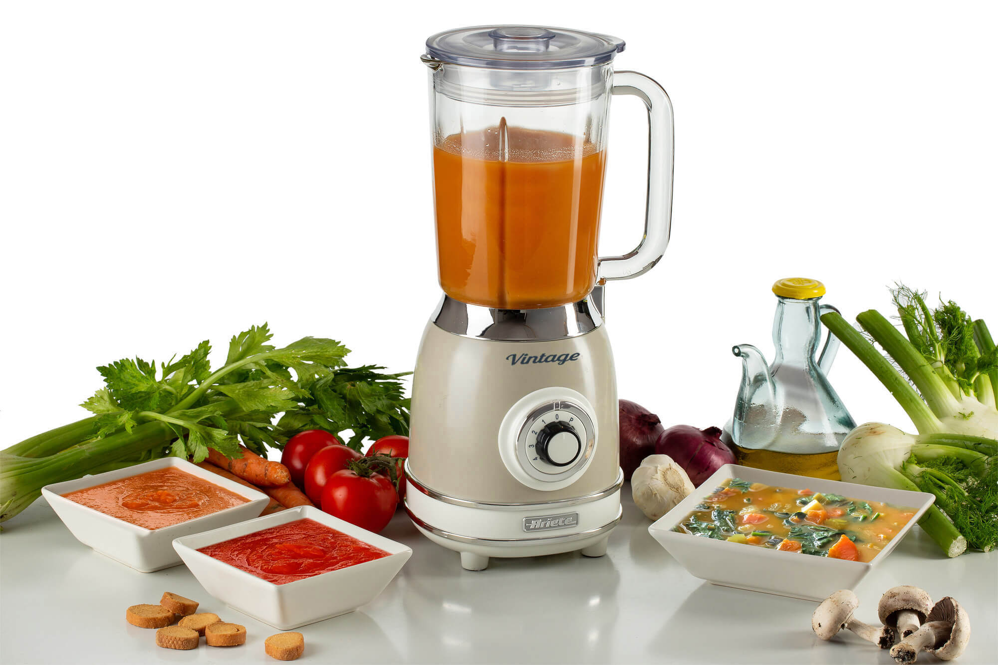 Turuncu sıvı içeren vintage blender, sebzeler ve çorba kaseleriyle çevrili.