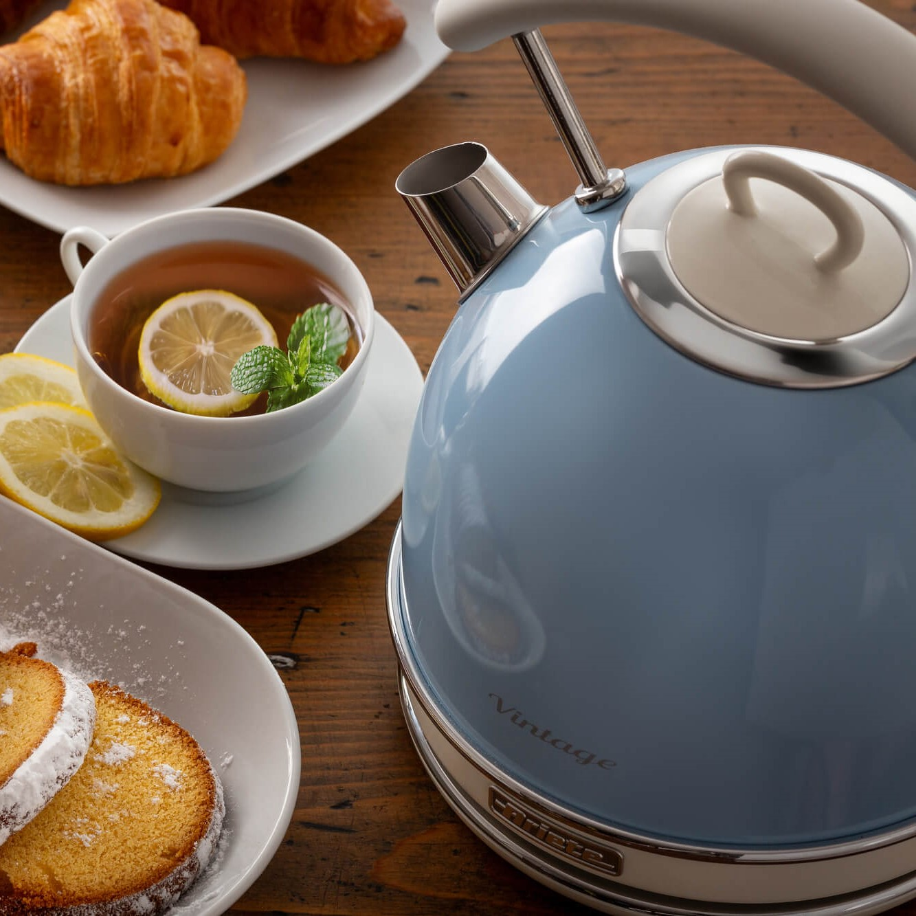 Een blauwe waterkoker met thee en citroen, croissants en cake op een tafel.