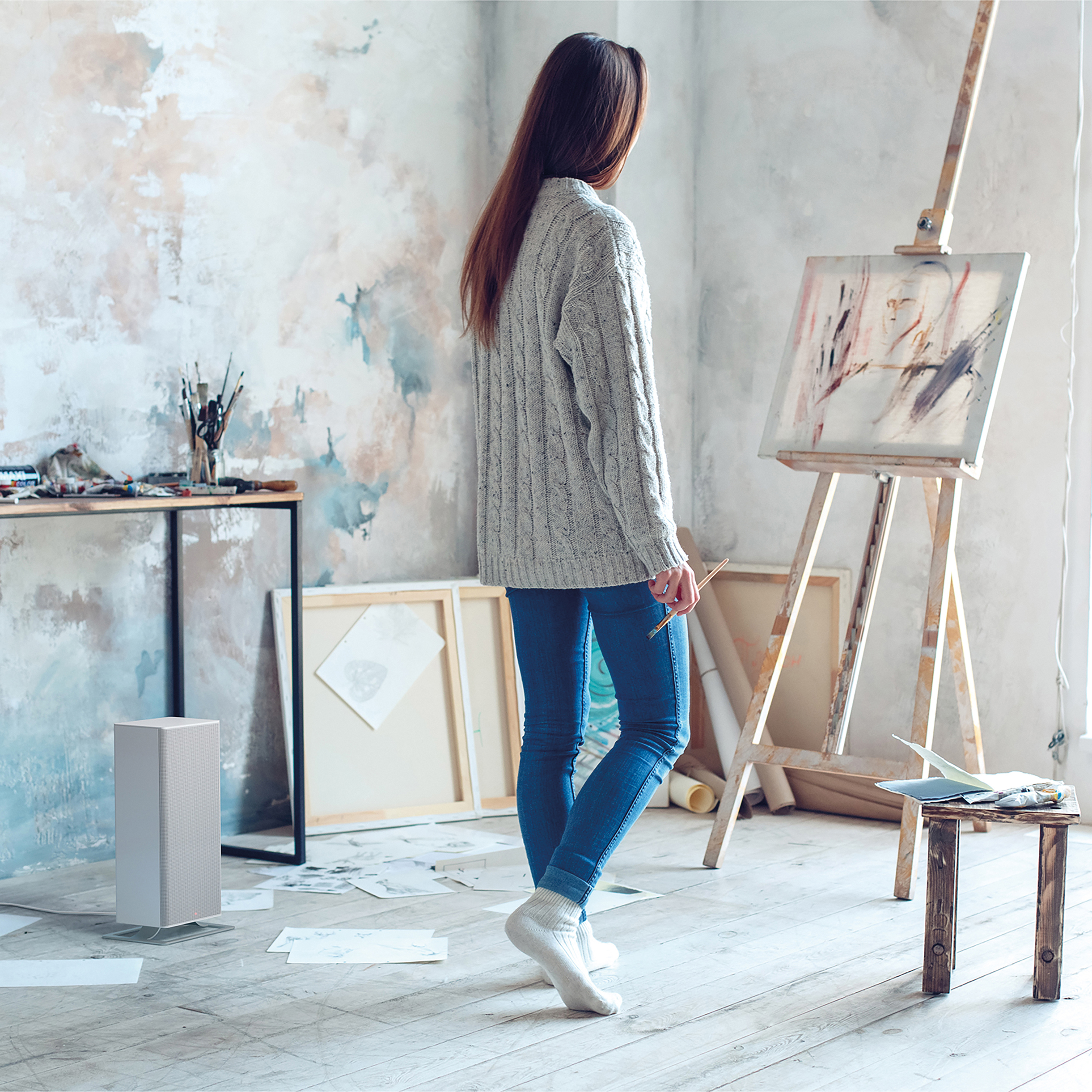 Een vrouw in een kamer kijkt naar een schilderij op een ezel. Een tafeltje en een kachel.
