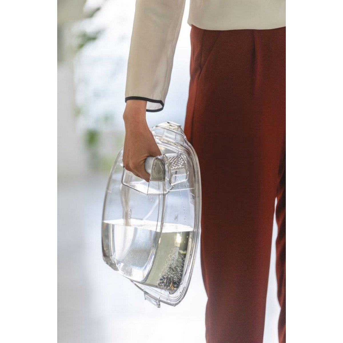 Una persona che tiene una brocca trasparente piena d'acqua, indossa una camicia bianca e pantaloni marroni.