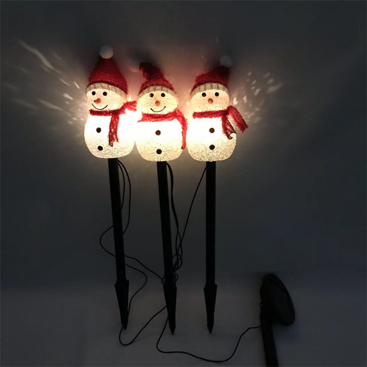 Drei beleuchtete Schneemann-Dekorationen mit roten Hüten und Schals, auf schwarzen Stäben, vor dunklem Hintergrund.
