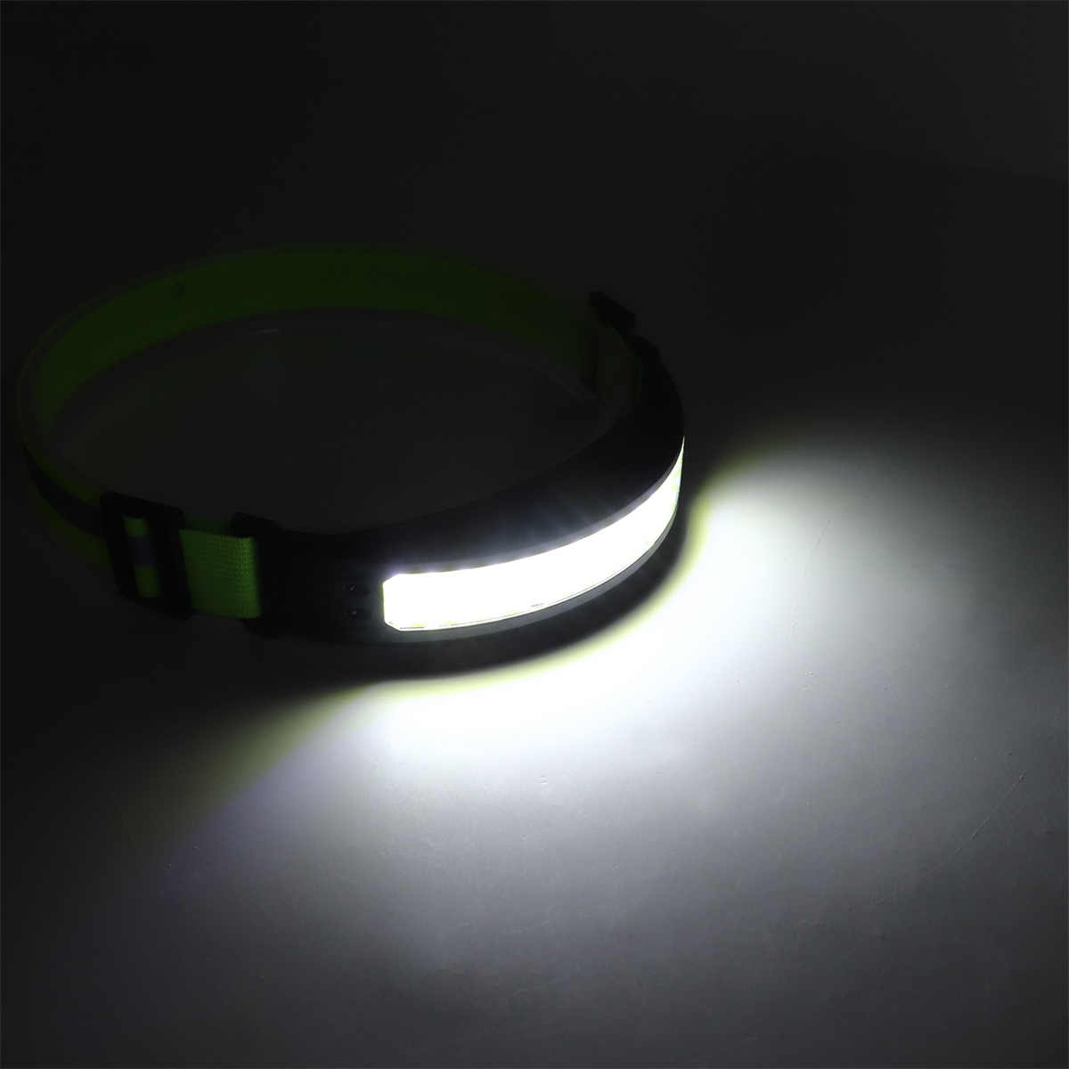 Stirnlampe mit hellem weißen Licht auf dunkler Oberfläche. Grünes Band, schwarzes Leuchtgehäuse.
